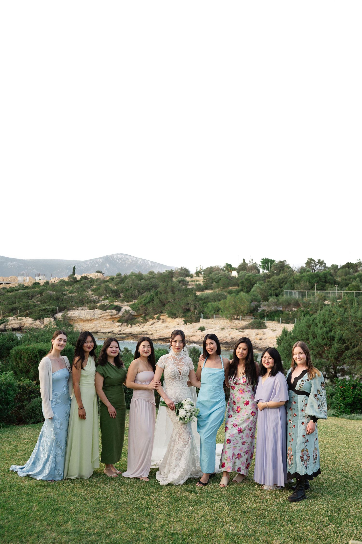Bride in lace gown poses with eight bridesmaids in pastel dresses on green lawn with mountain backdrop.