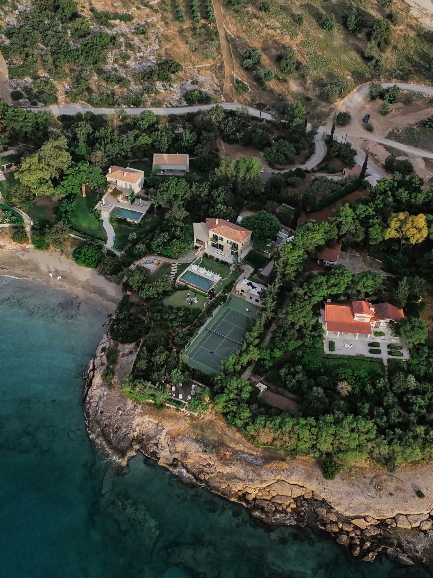 Aerial view of a luxury coastal estate surrounded by lush greenery, tennis court, and turquoise Mediterranean waters.