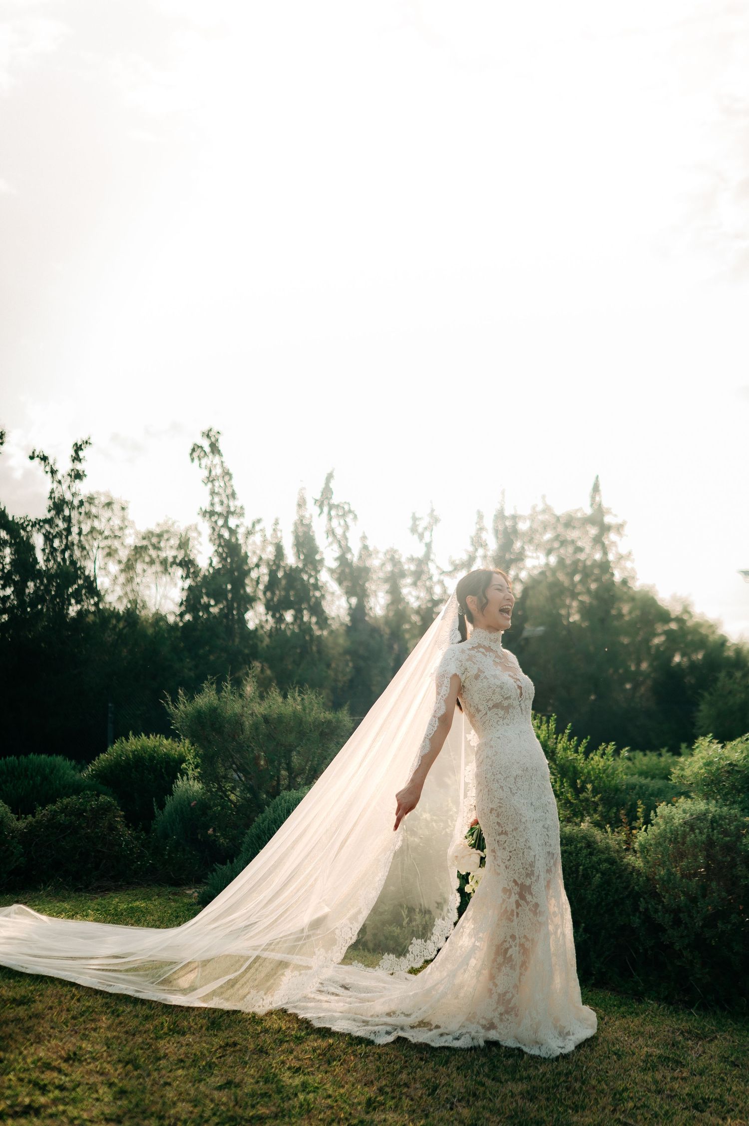Bride in fitted lace gown with long cathedral veil posing gracefully in sunlit green garden at dusk.