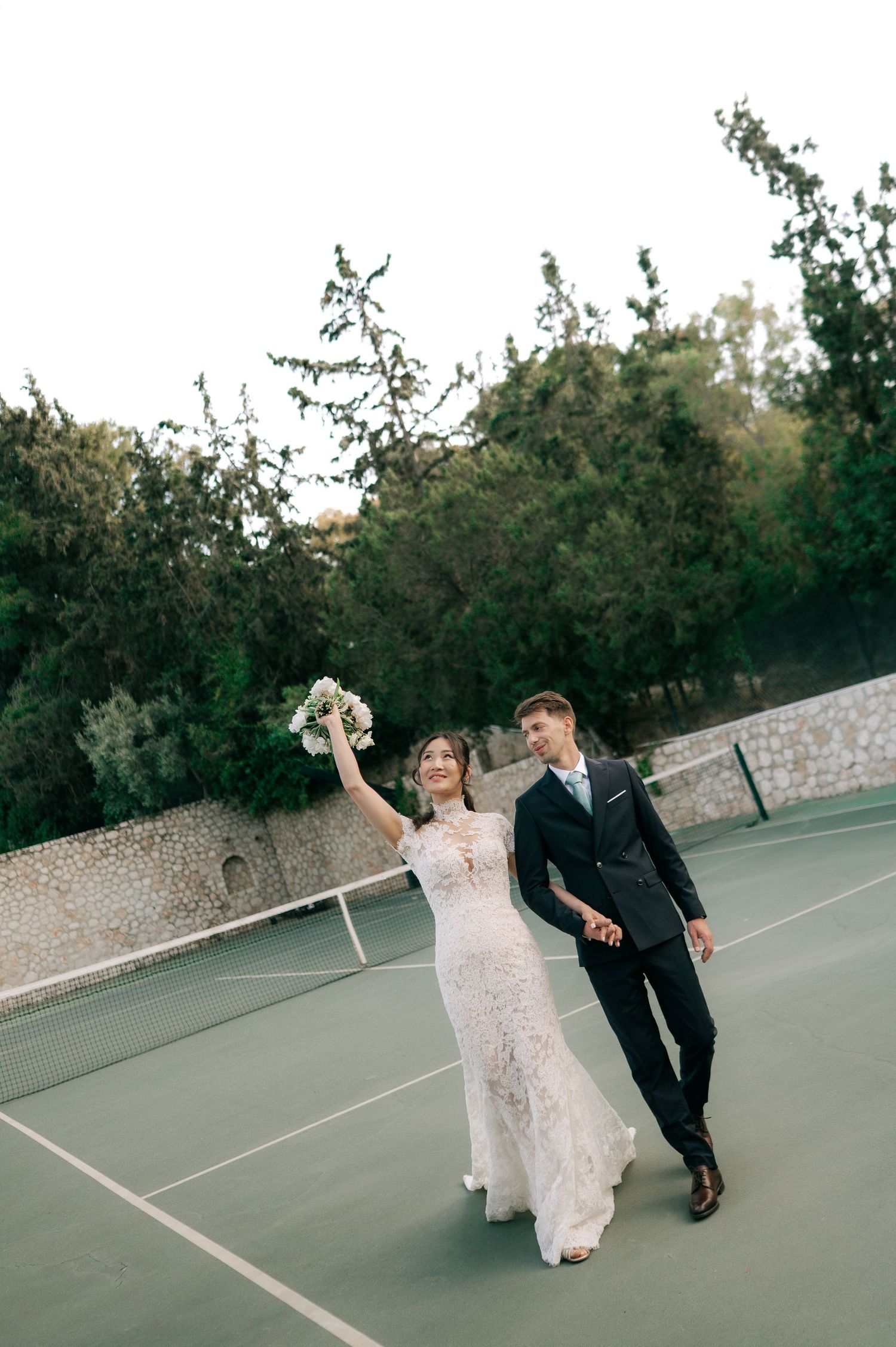 Bride in lace gown raises bouquet joyfully while walking hand-in-hand with groom on a tennis court.