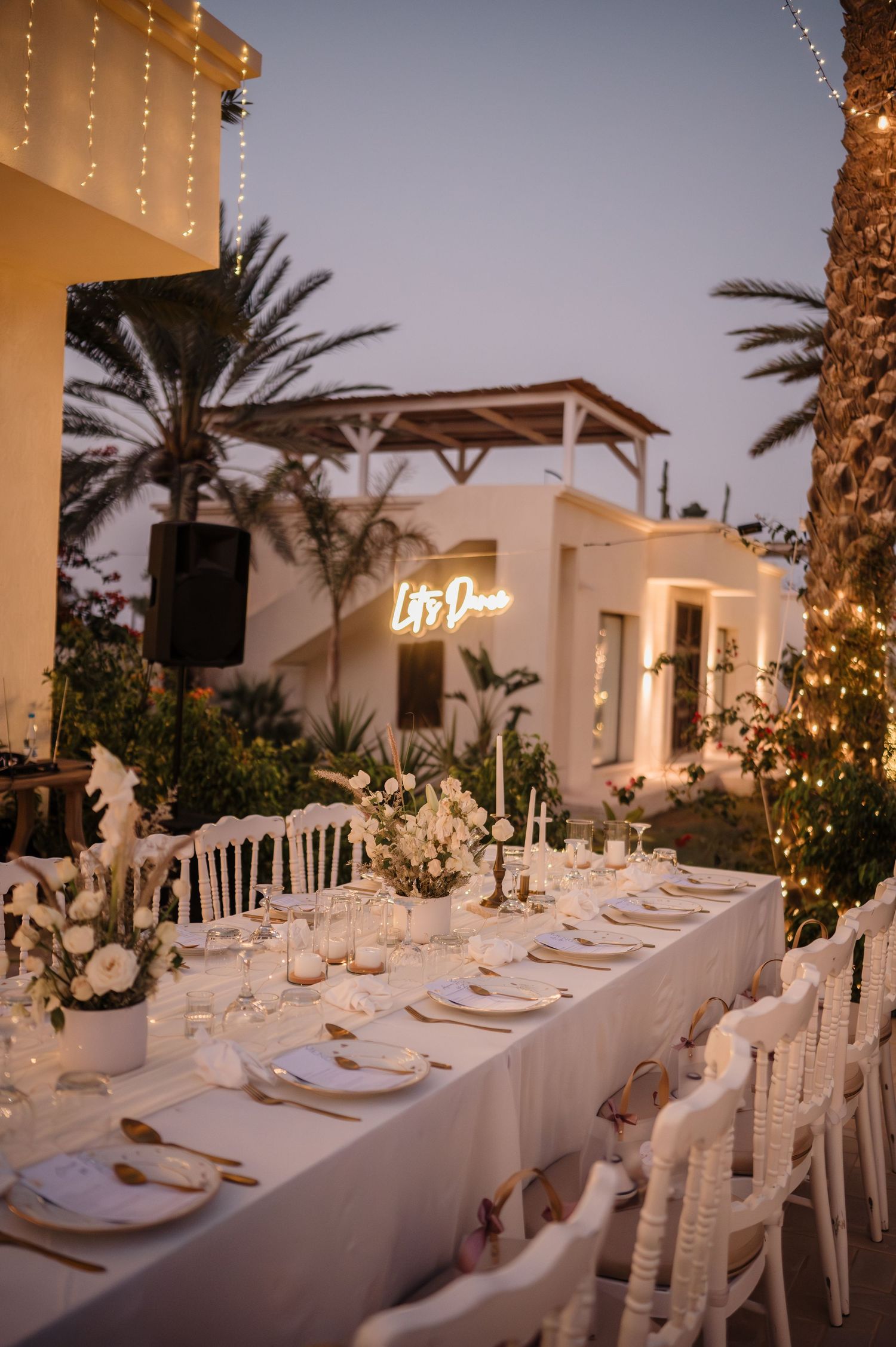 Romantic outdoor wedding reception at twilight with long white table, candles, and glowing neon sign decor.