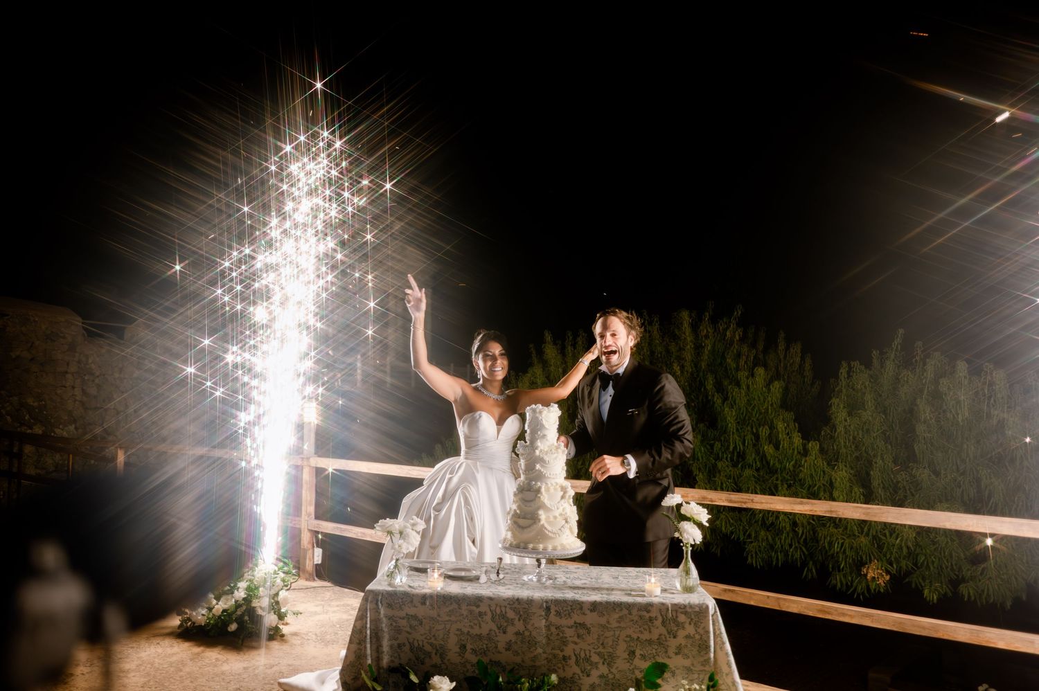 Joyful newlyweds celebrate beside towering wedding cake with dazzling firework sparklers at night.