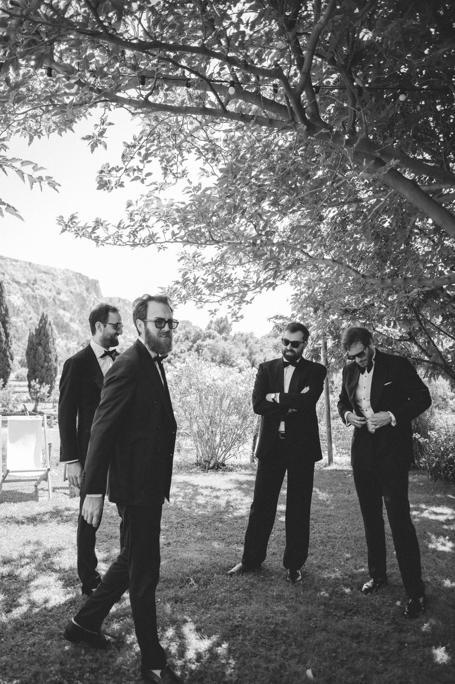 Black and white photo of four groomsmen in tuxedos relaxing under a tree in a garden setting.