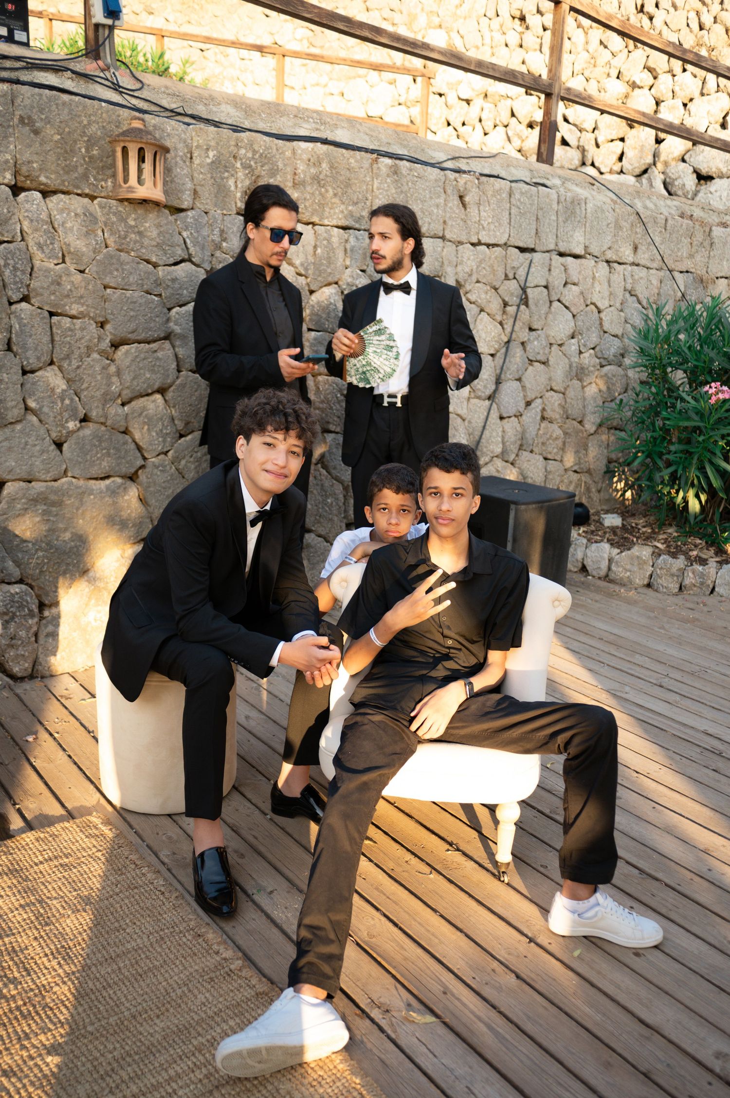 Group of young male wedding guests in black tuxedos posing on wooden terrace with stone wall backdrop.