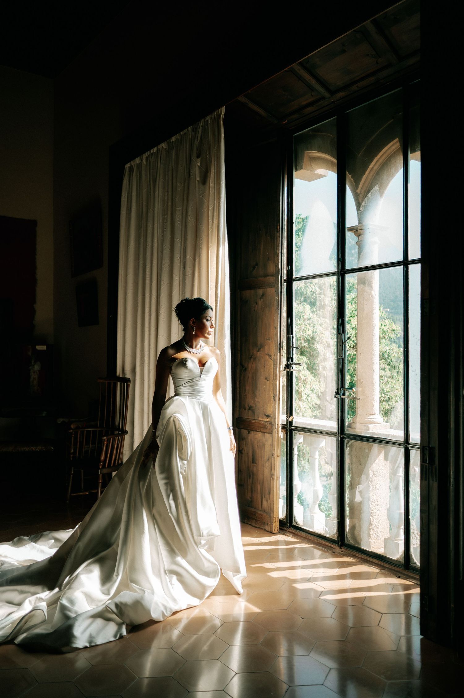 Bride in flowing white ball gown stands by ornate arched windows with sunlight streaming in.