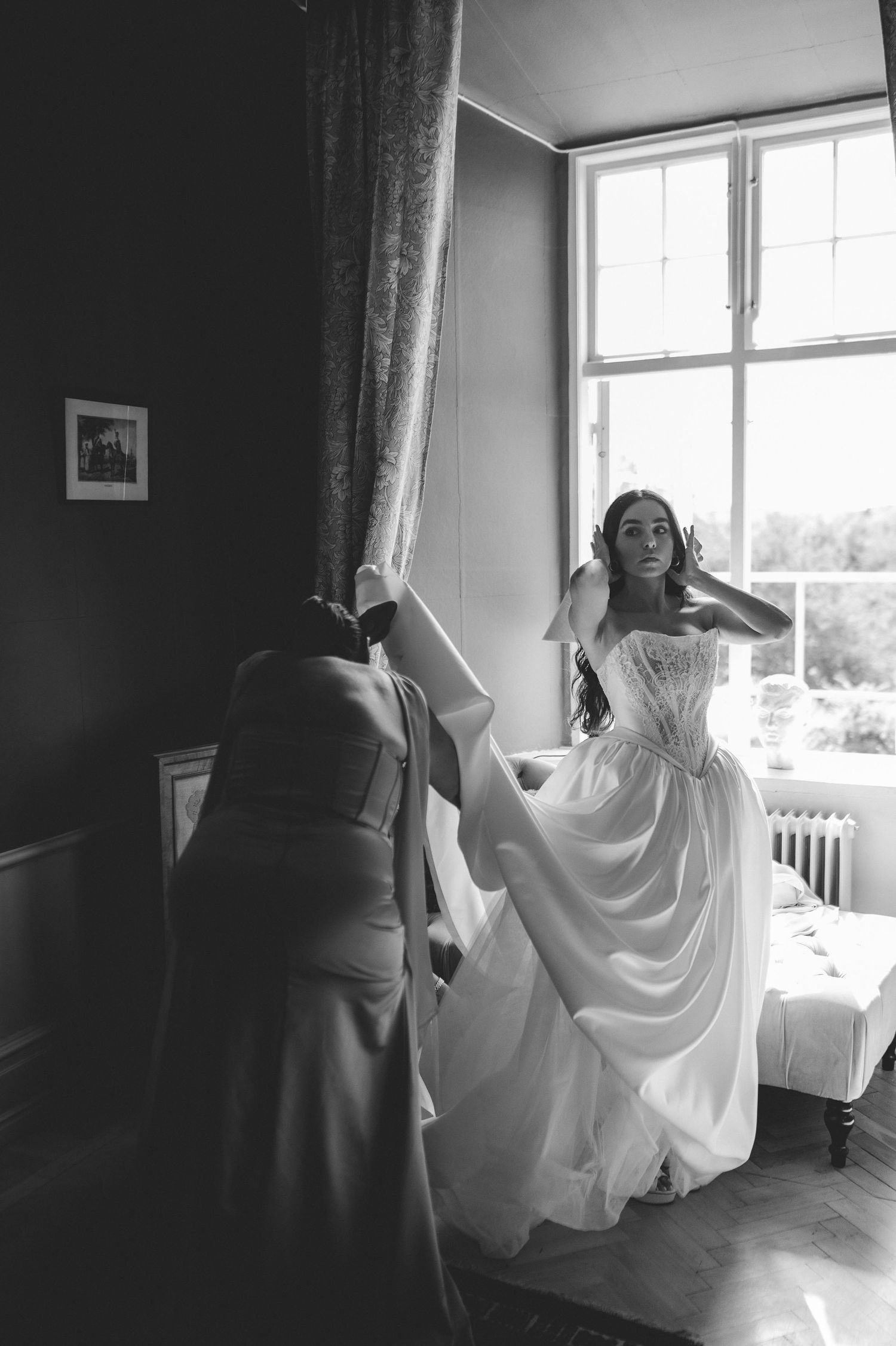 Black and white photo of bride adjusting veil near window with bridesmaid holding dress.