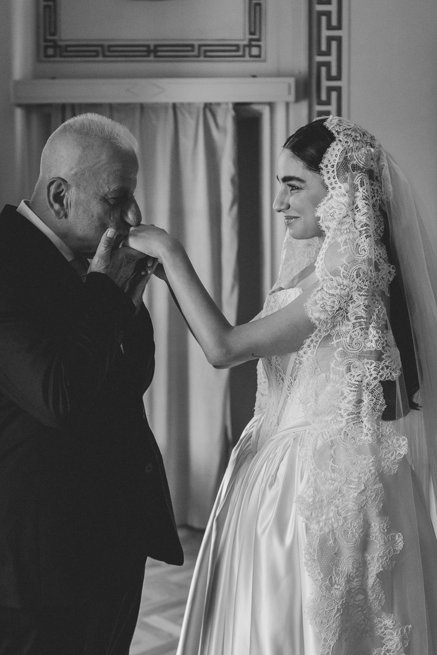 Black and white photo of emotional father kissing bride's hand on her wedding day in ornate room.