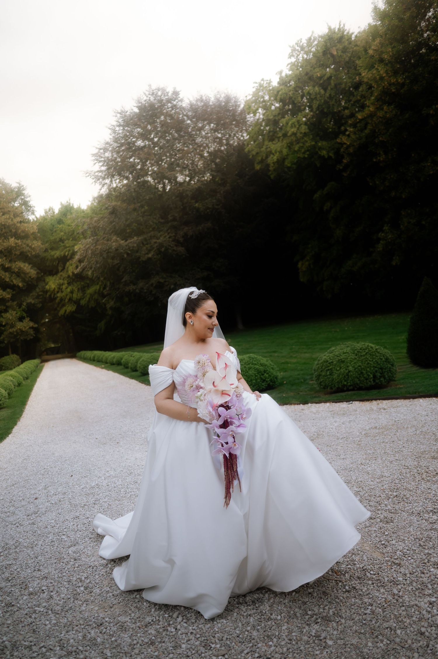 Elegant bride in off-shoulder ball gown holding cascading pink orchid bouquet on tree-lined garden path at dusk.
