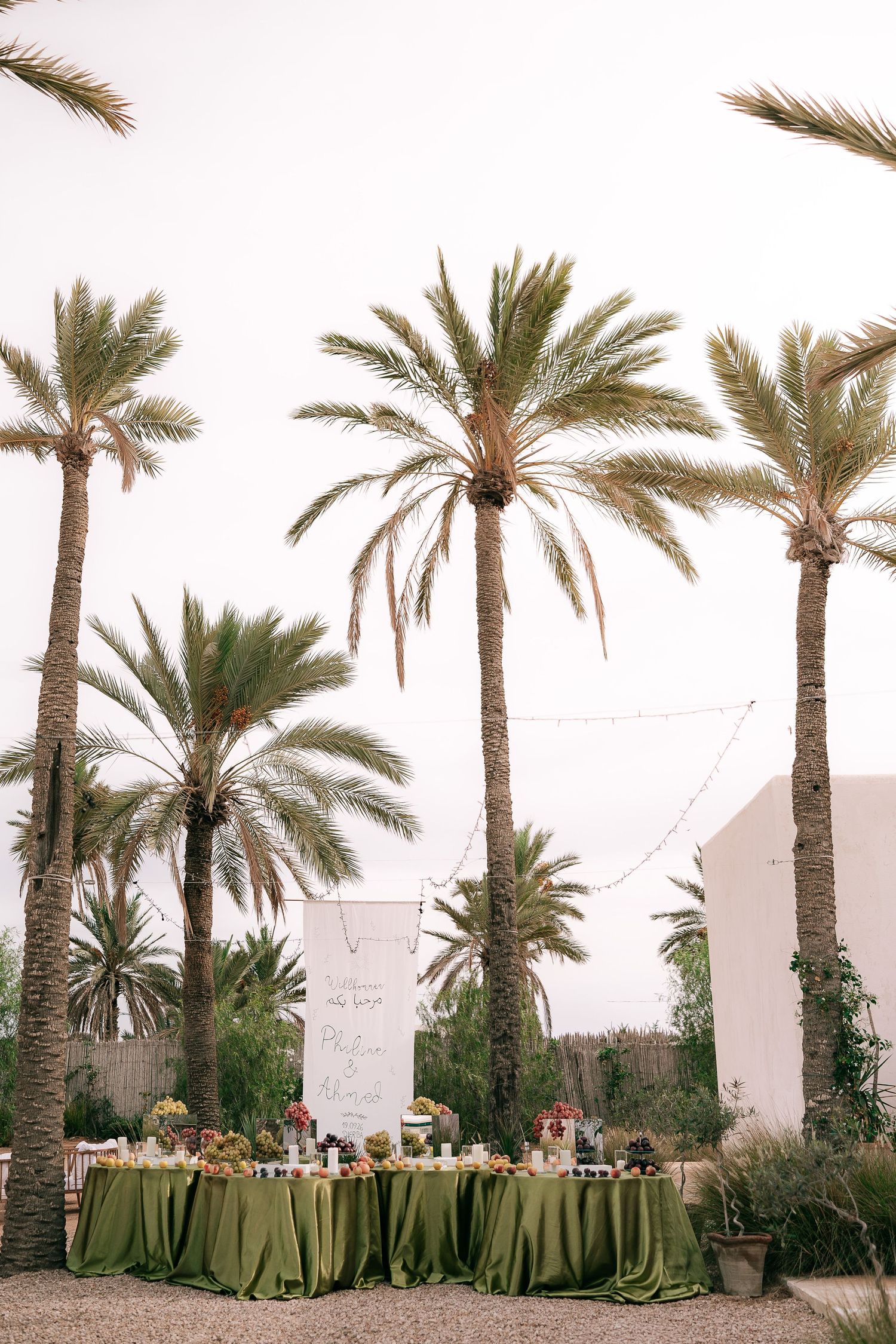 Elegant outdoor wedding reception tables with green linens set beneath tall palm trees.