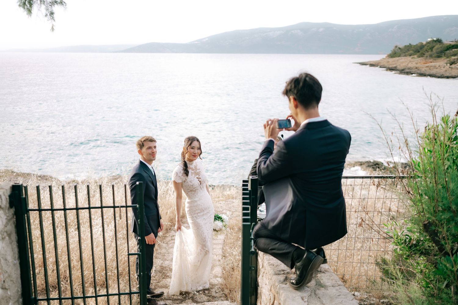Photographer capturing bride and groom portrait near iron gate overlooking scenic blue sea.