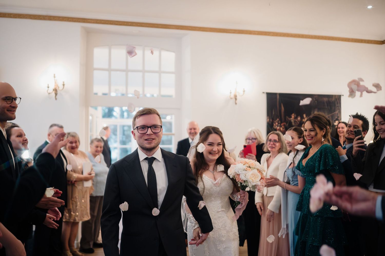 Smiling bride and groom walk hand in hand through applauding guests in bright wedding venue.