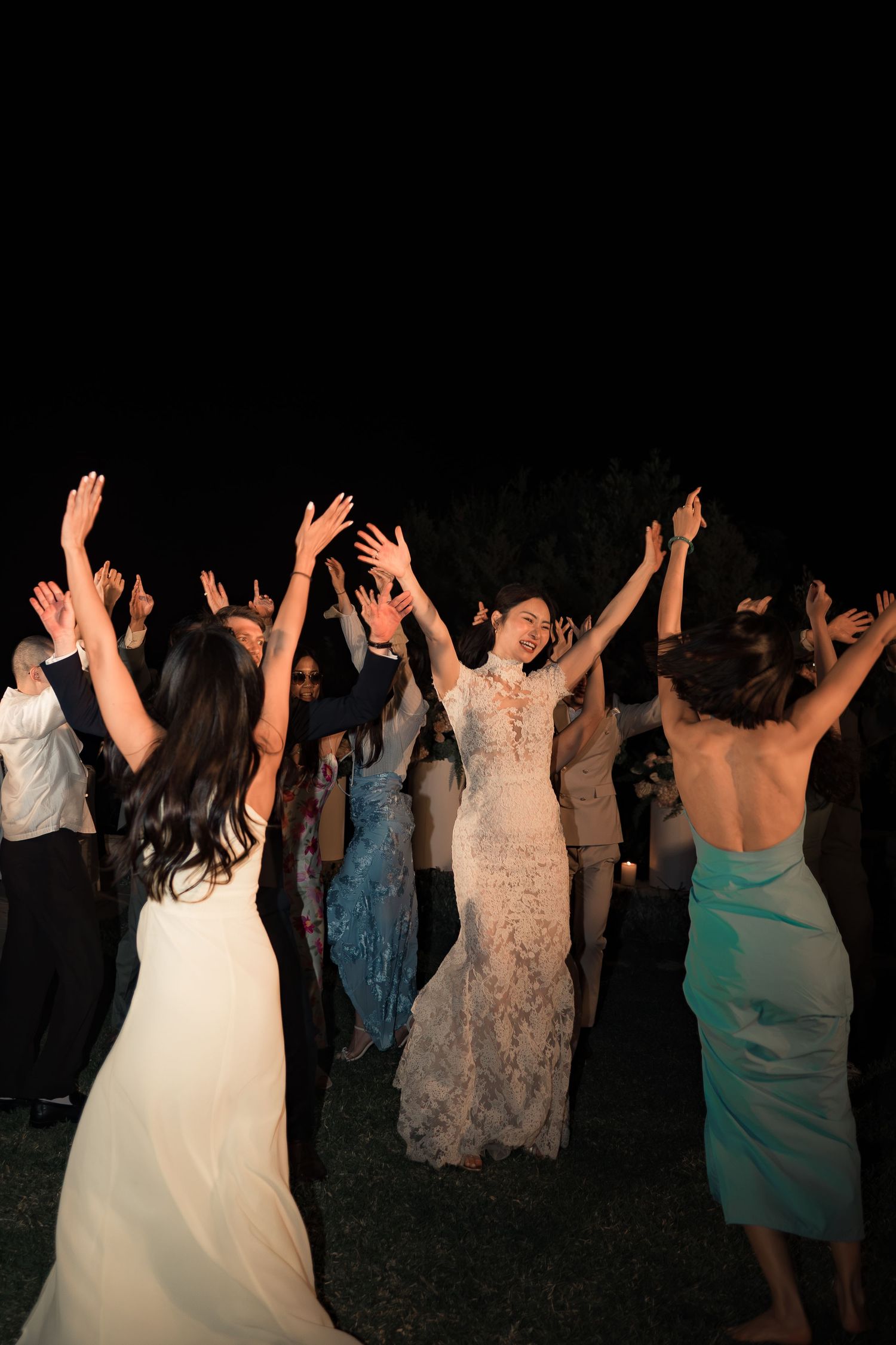 Bride in lace gown dances joyfully with guests, arms raised, at an outdoor nighttime wedding reception.
