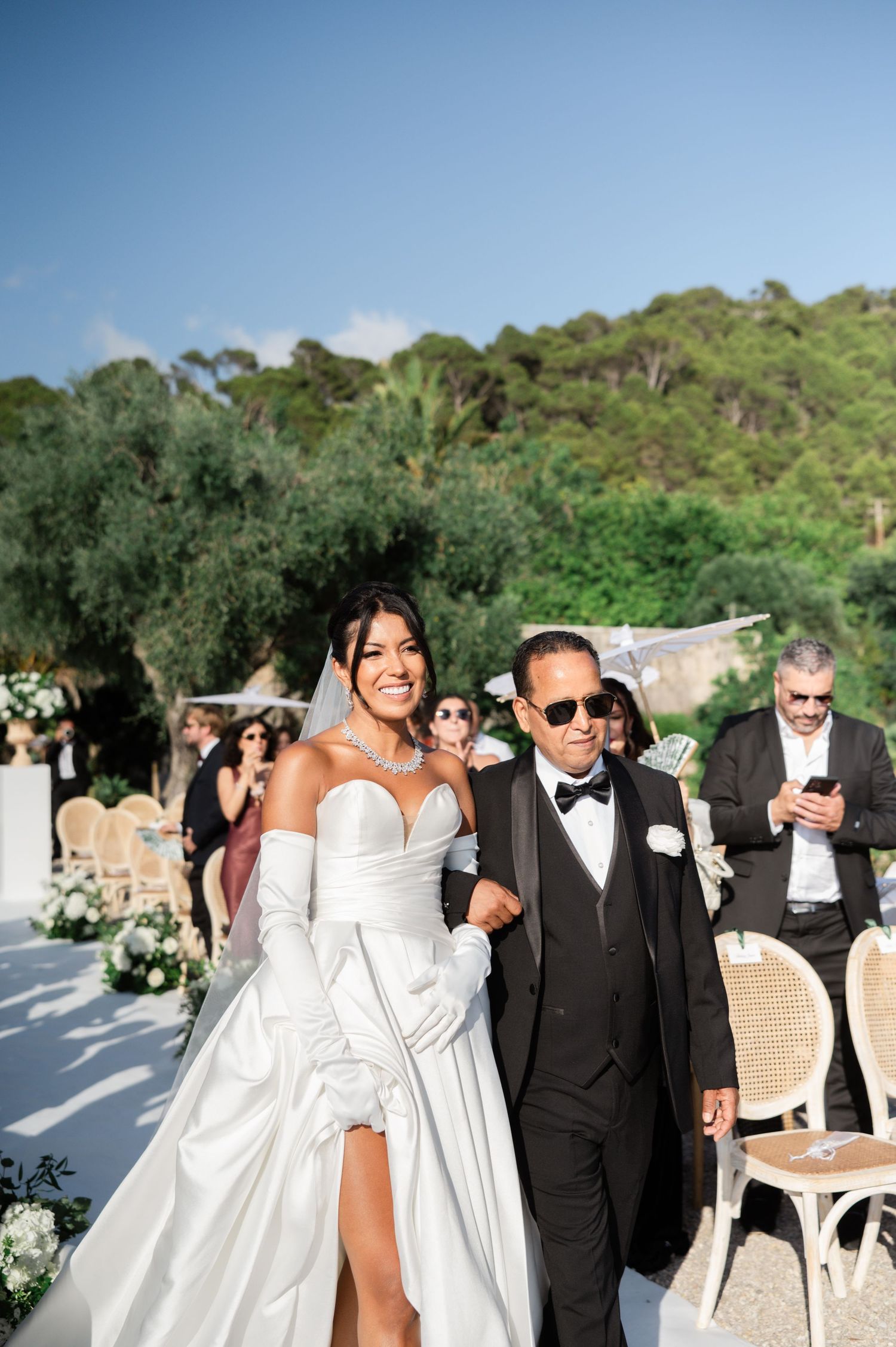 Radiant bride in a strapless white gown with gloves walks down the aisle with her father.