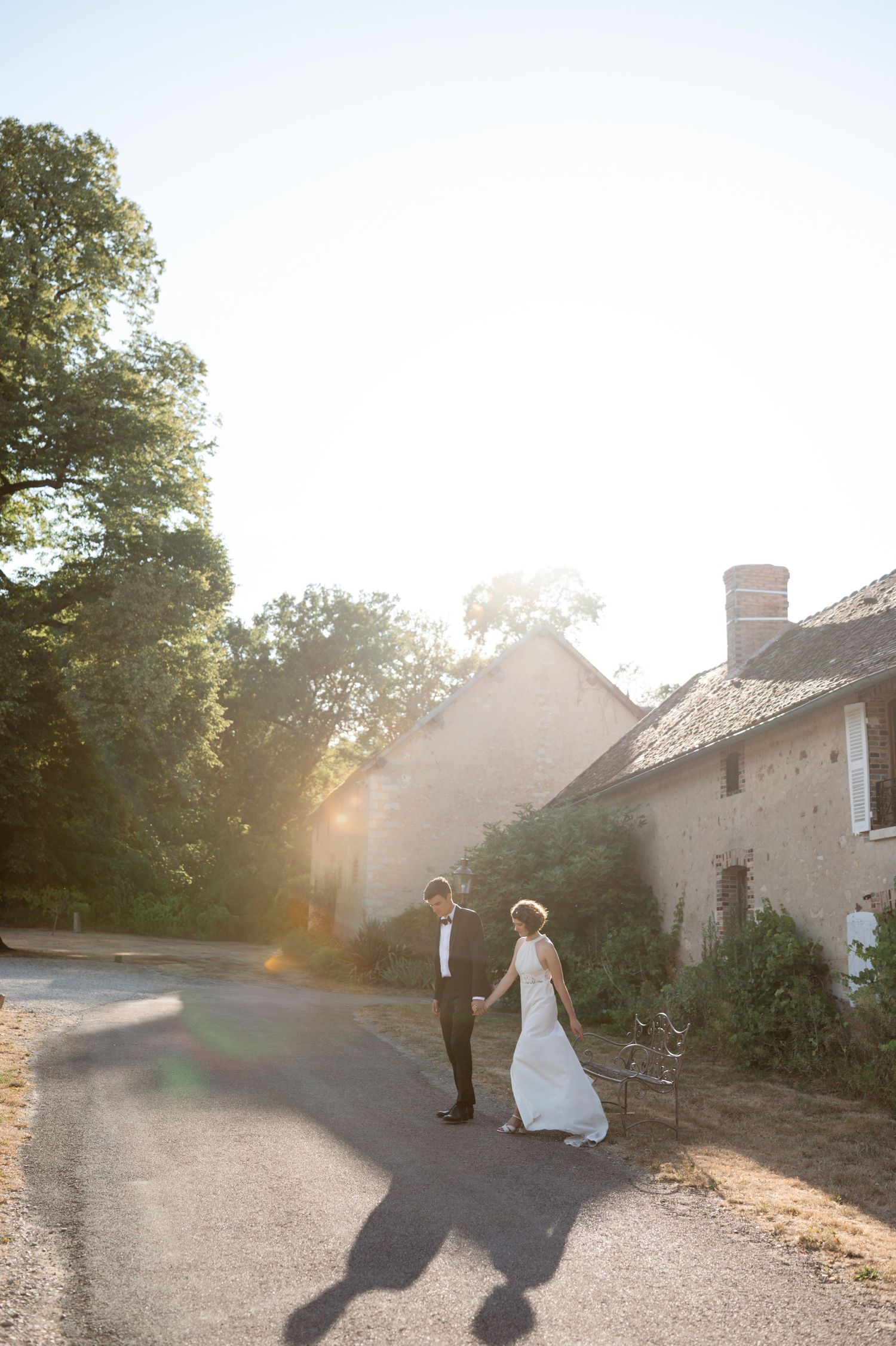 Golden hour wedding portrait of bride and groom strolling past rustic stone buildings with beautiful sun flare backdrop.