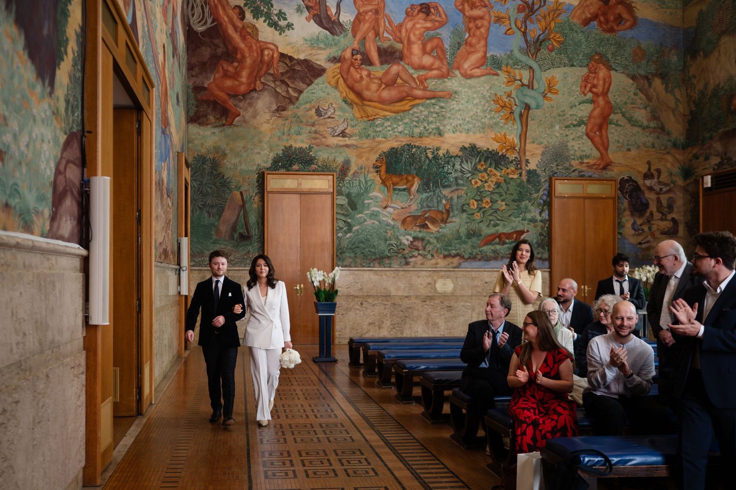 Couple walks down aisle in ornate hall with colorful murals as guests applaud during elegant wedding ceremony.