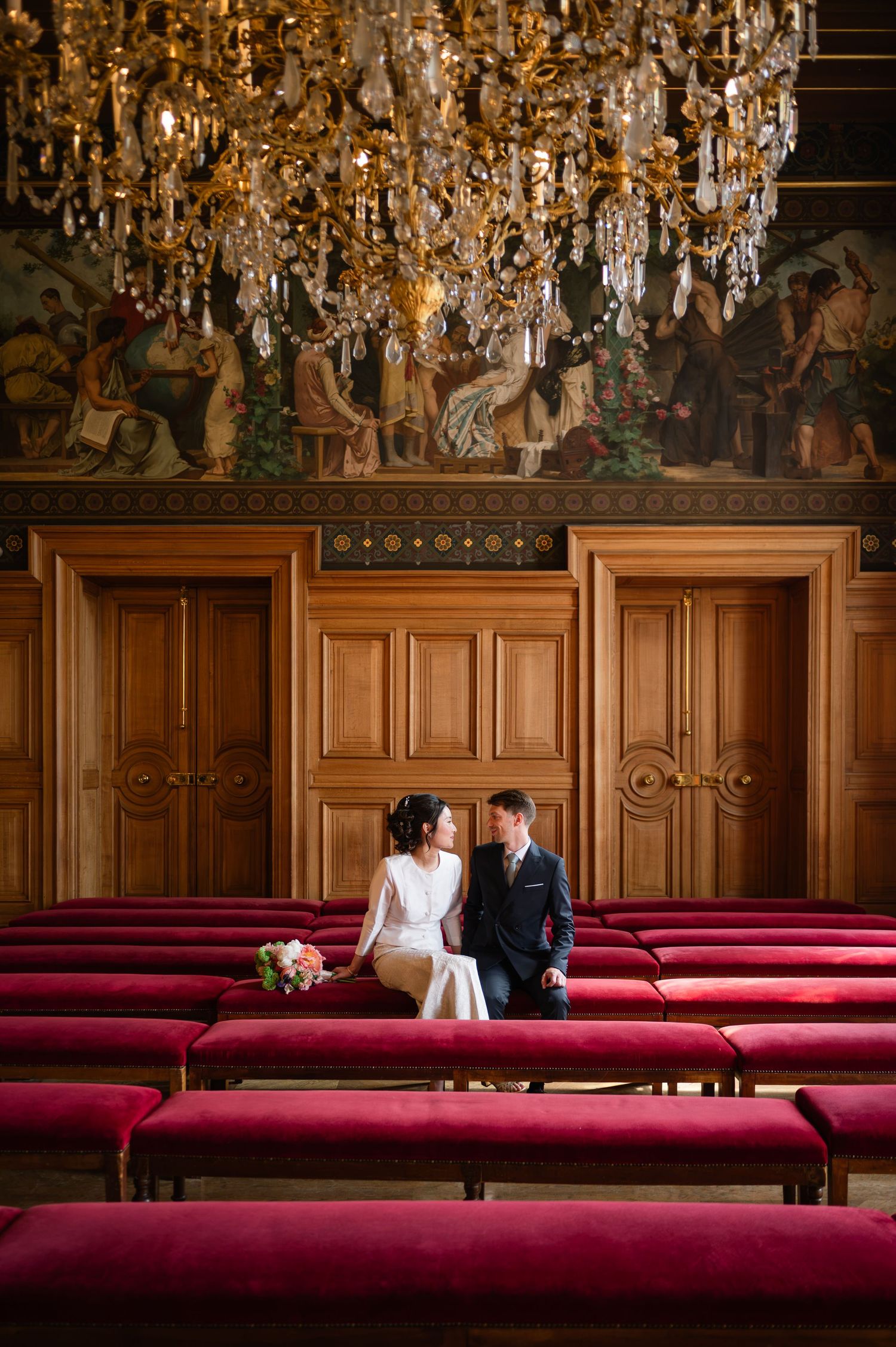 Bride and groom sitting together in an ornate hall with red velvet benches and a grand crystal chandelier above.