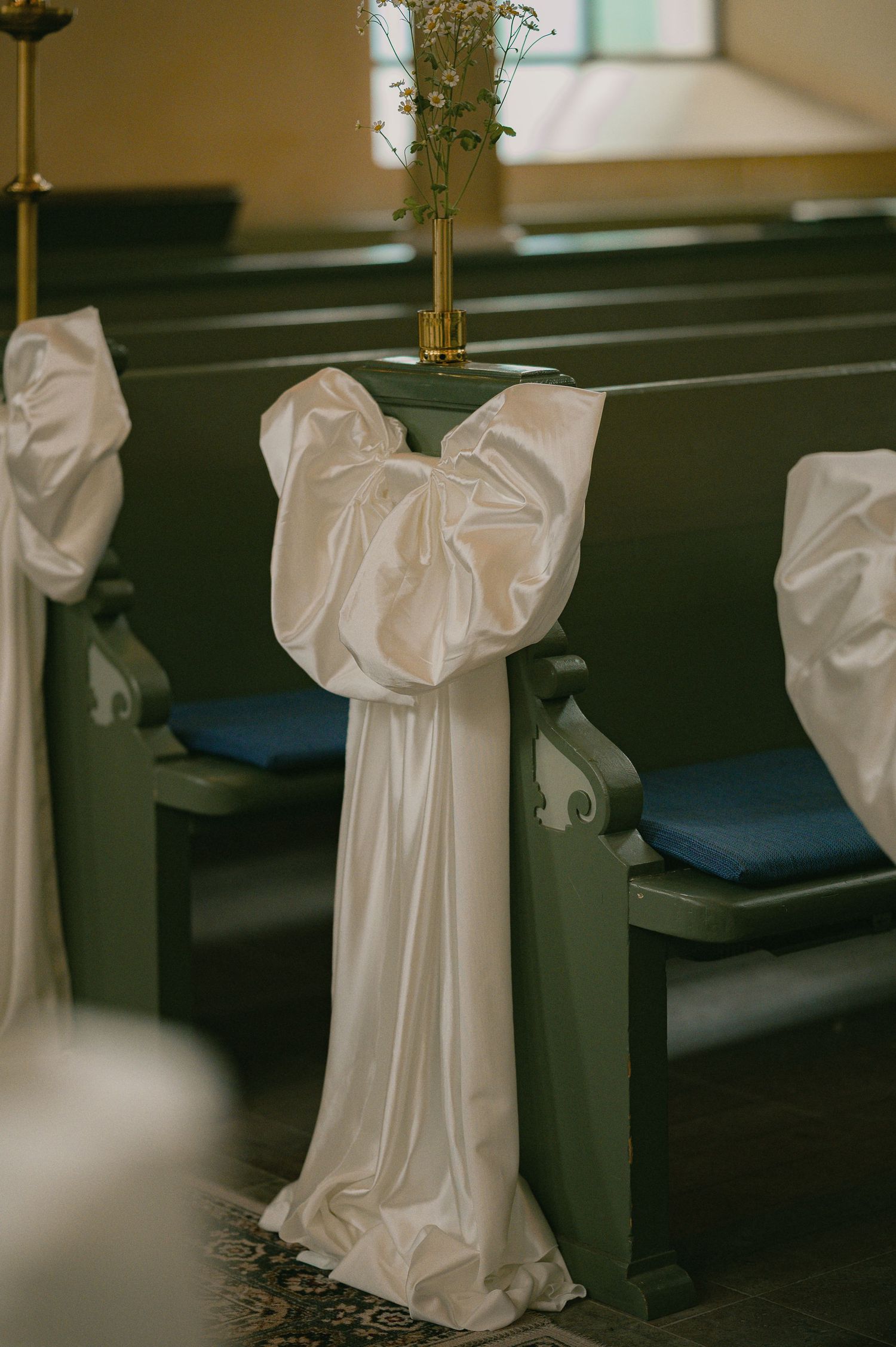 White satin bow decoration on dark green wooden church pew with floral arrangement at wedding ceremony.
