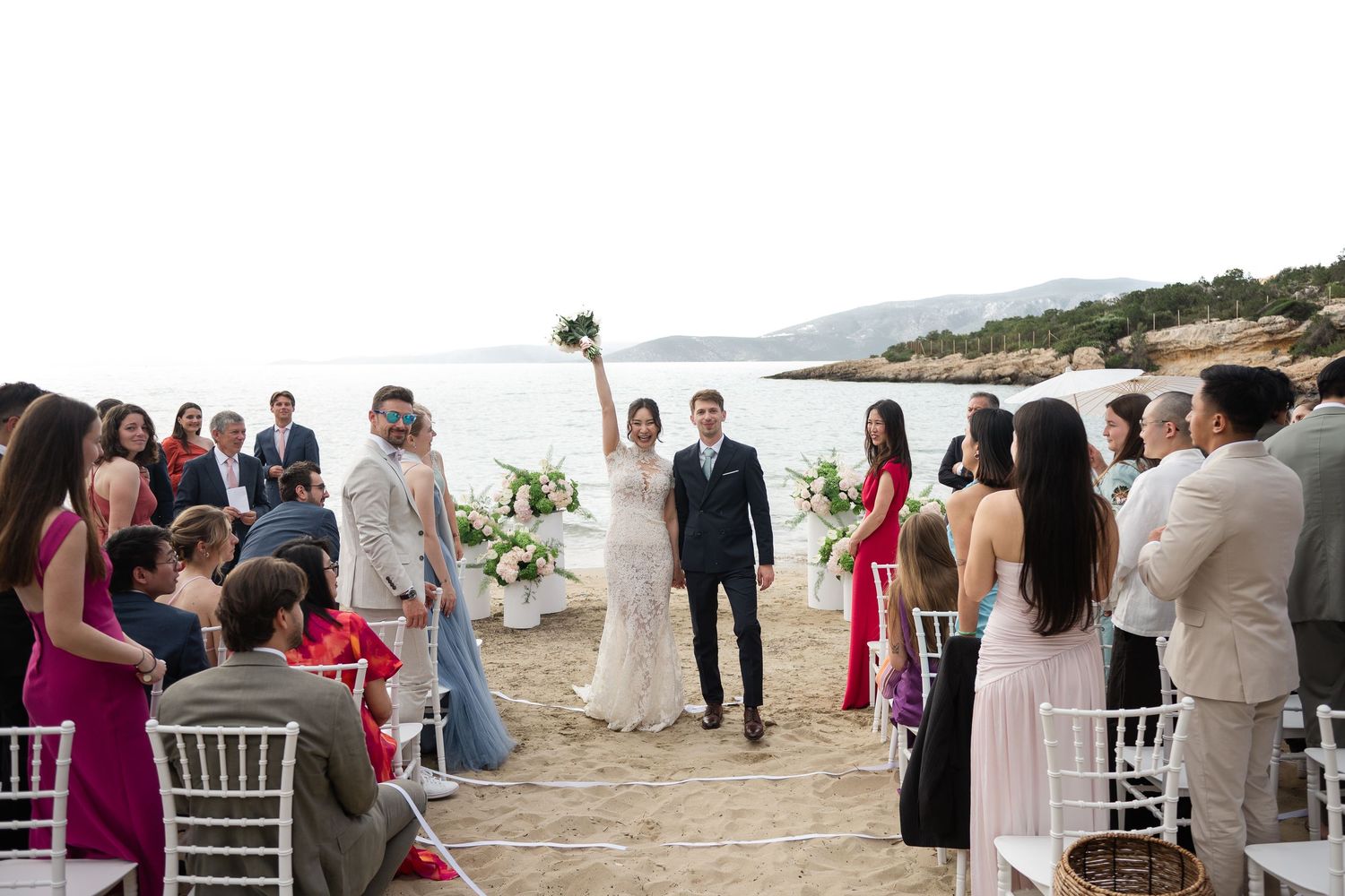 Newlyweds celebrate with raised bouquet as guests cheer at beach wedding ceremony.