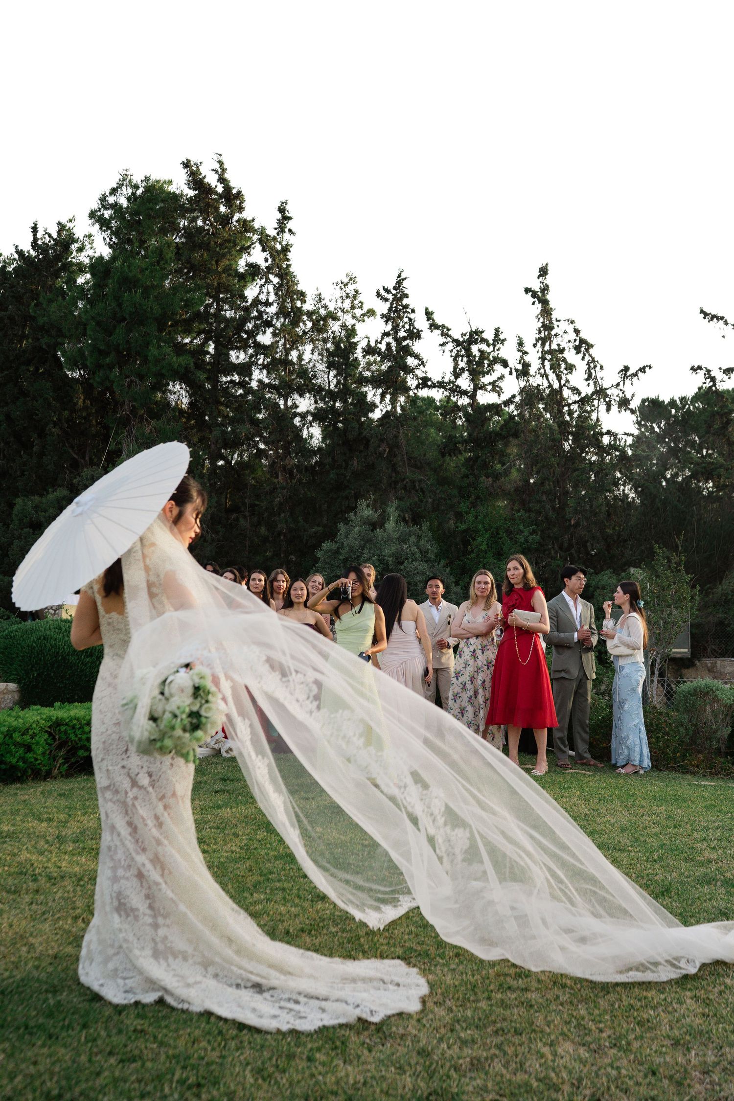 Bride in lace gown with parasol lets her veil flow in the breeze before wedding guests outdoors.
