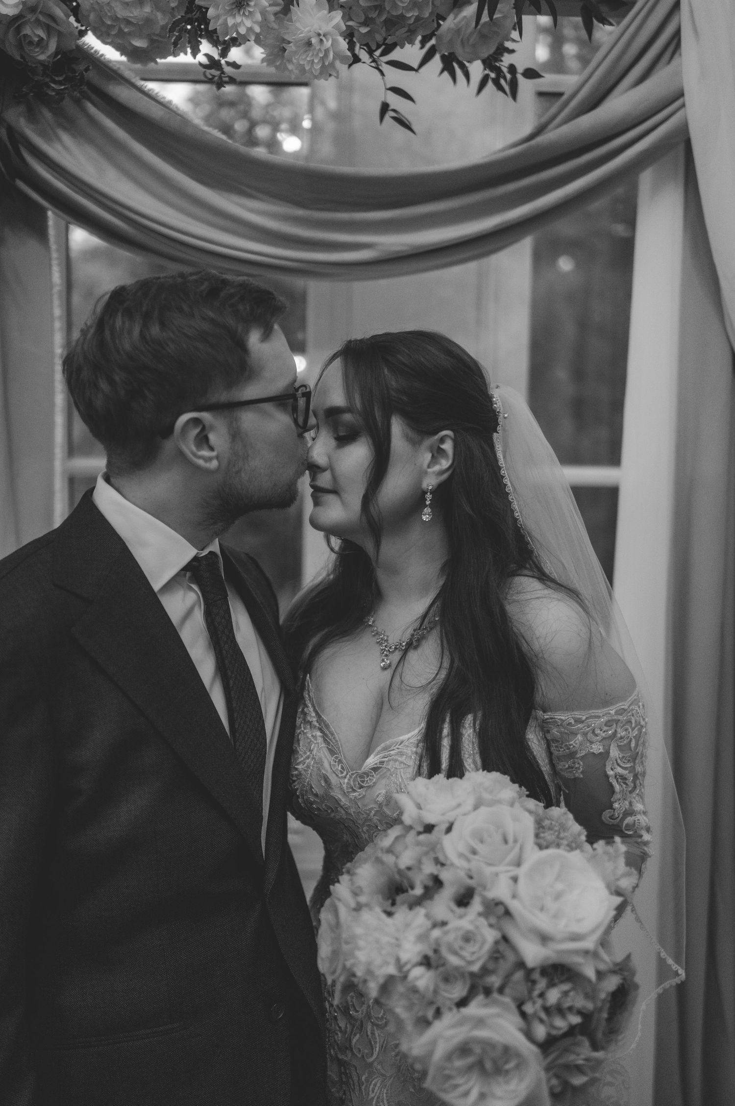 Black and white photo of bride and groom sharing a kiss under floral arch, bride holding white rose bouquet.