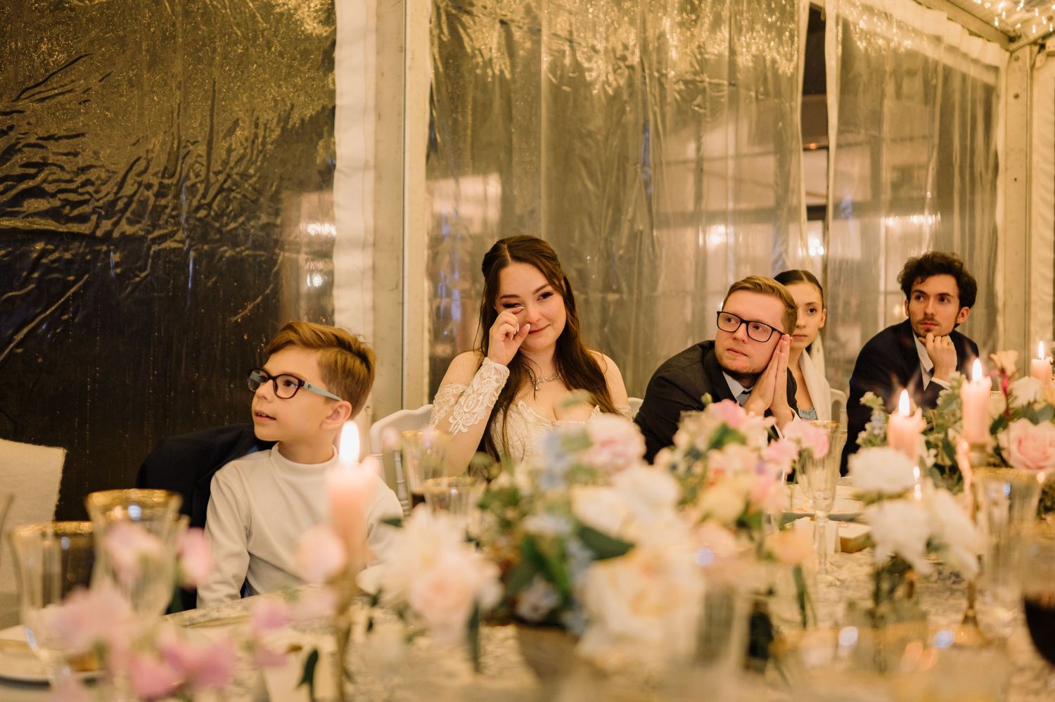 Wedding guests listening to emotional speeches at a candlelit reception table with pink floral centerpieces.