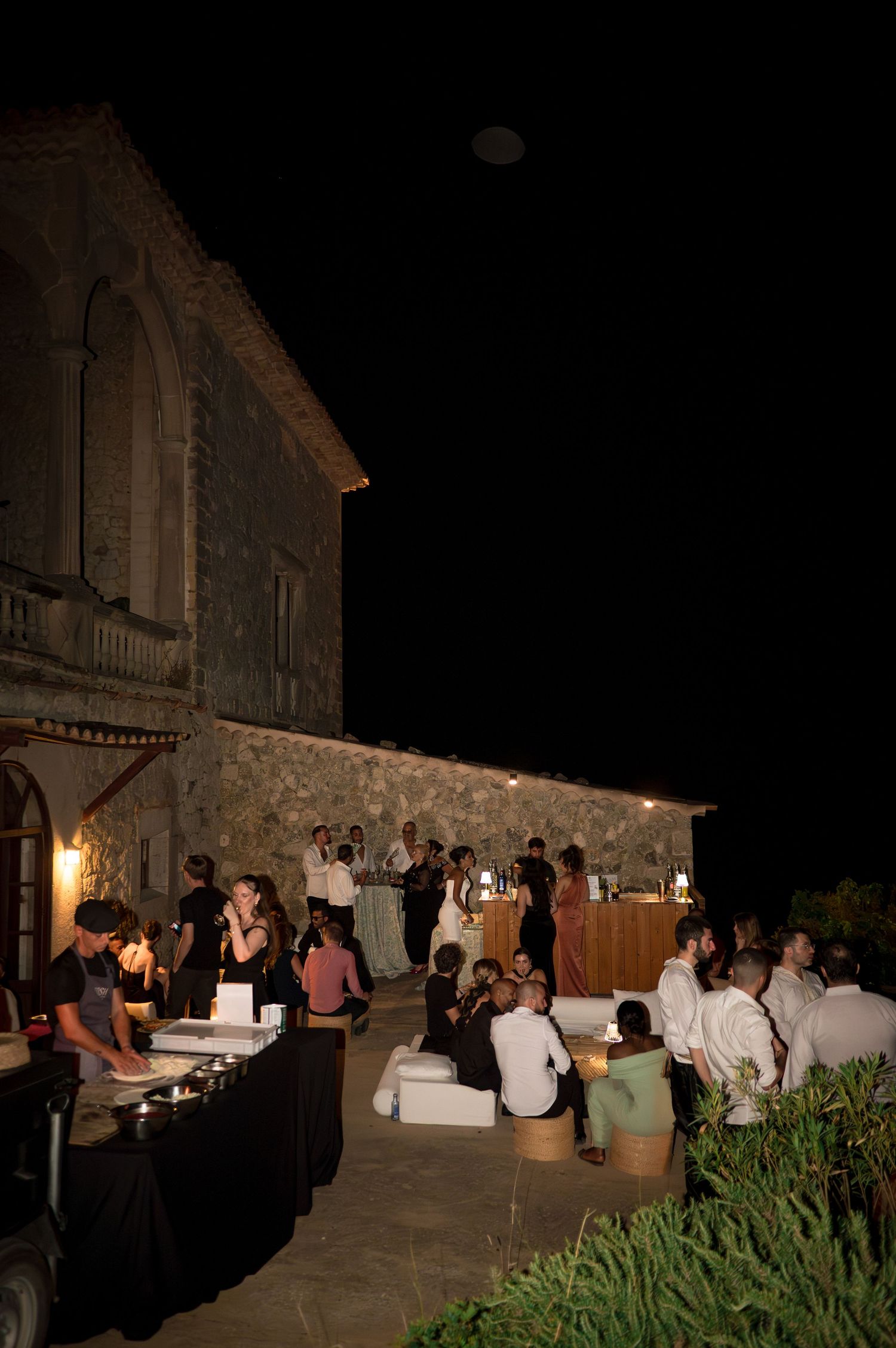 Guests mingling outdoors at night near a historic stone building at a wedding reception.