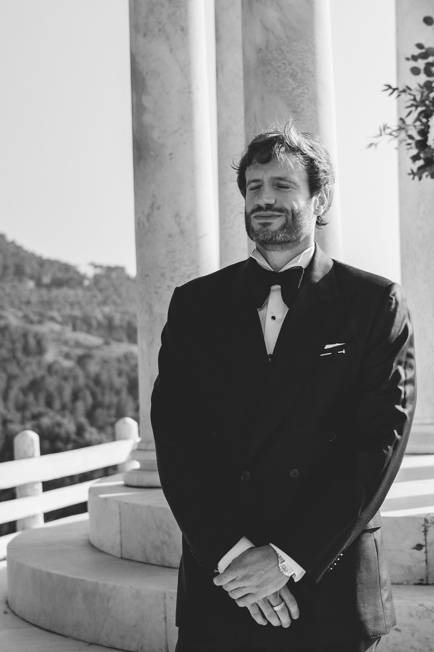 Black and white portrait of a bearded groom in a tuxedo standing between marble columns.