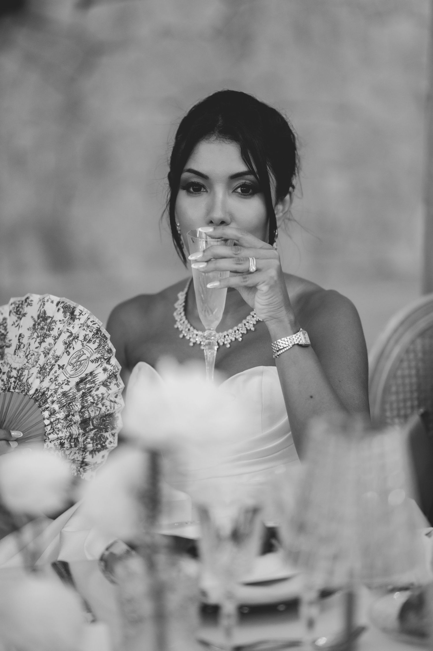 Beautiful bride holding decorative floral fan at wedding reception table wearing statement necklace.