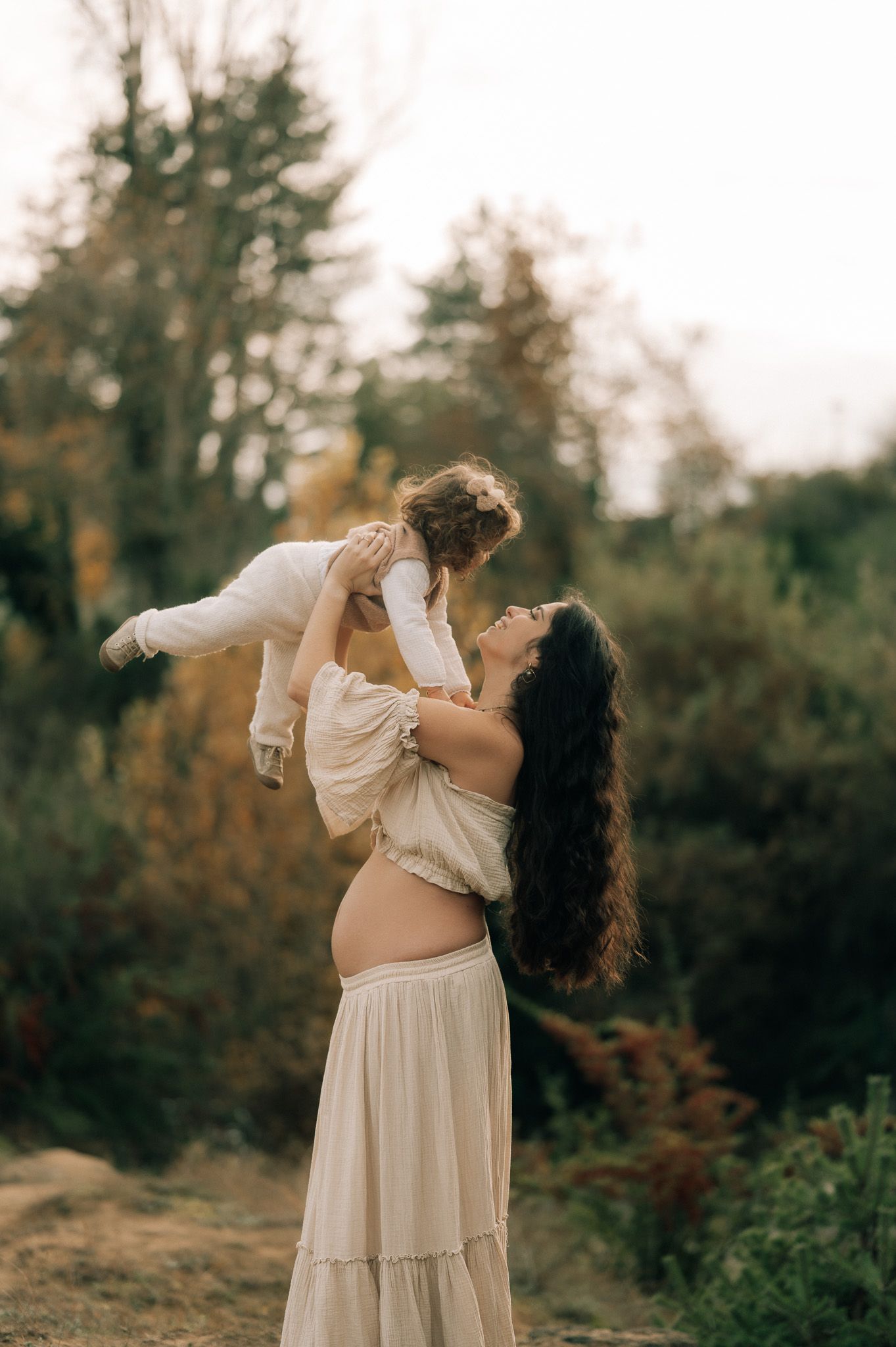 Pregnant woman in cream dress lifting toddler outdoors among autumn trees during a maternity photo session.