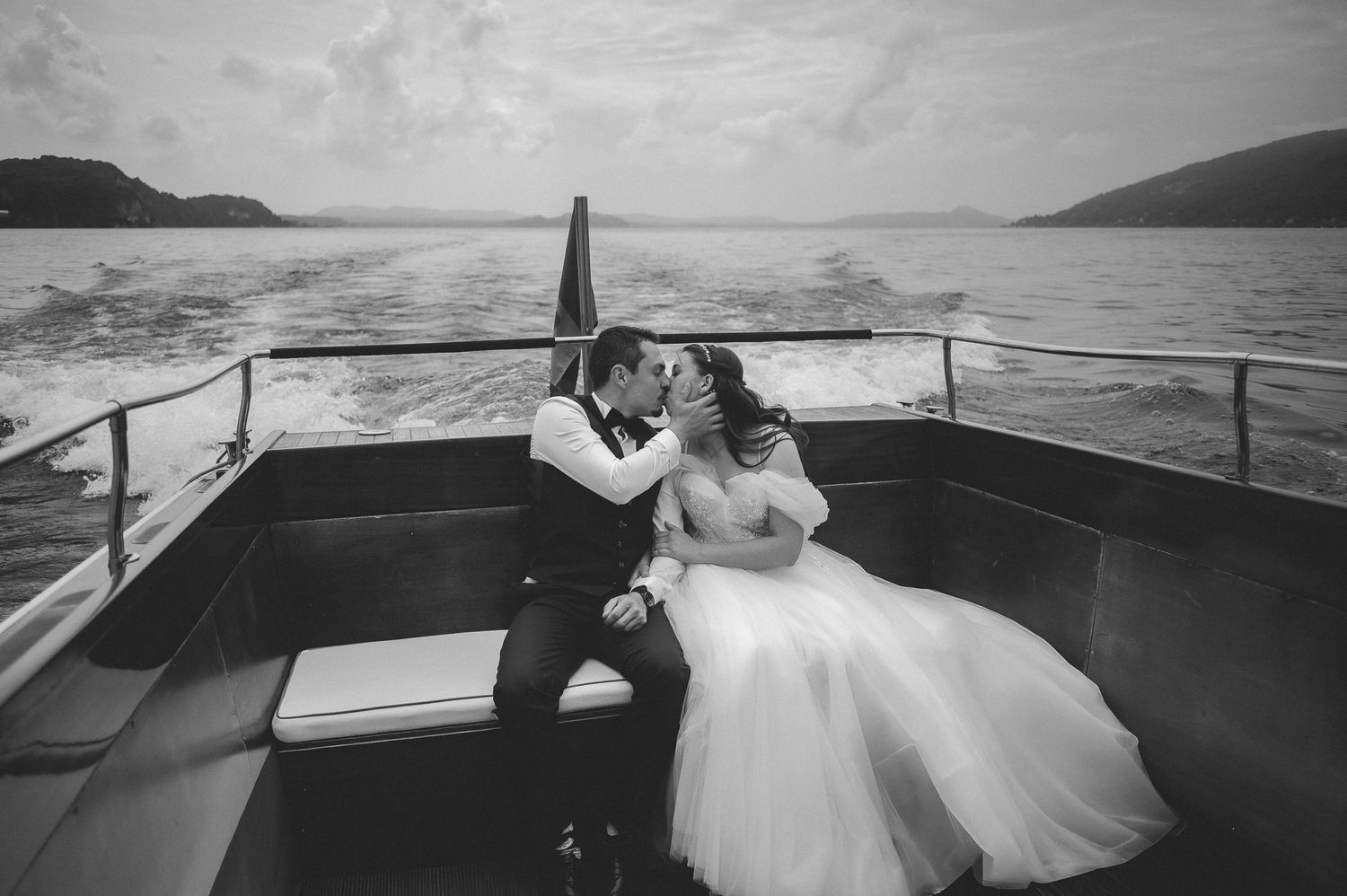Black and white photo of a bride and groom sharing a romantic kiss on a boat with scenic water and hills behind them.