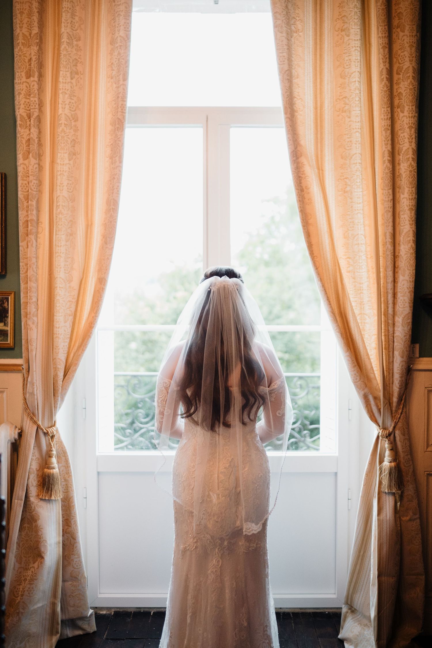 Bride in lace wedding gown and veil viewed from behind gazing out a large window with gold curtains.