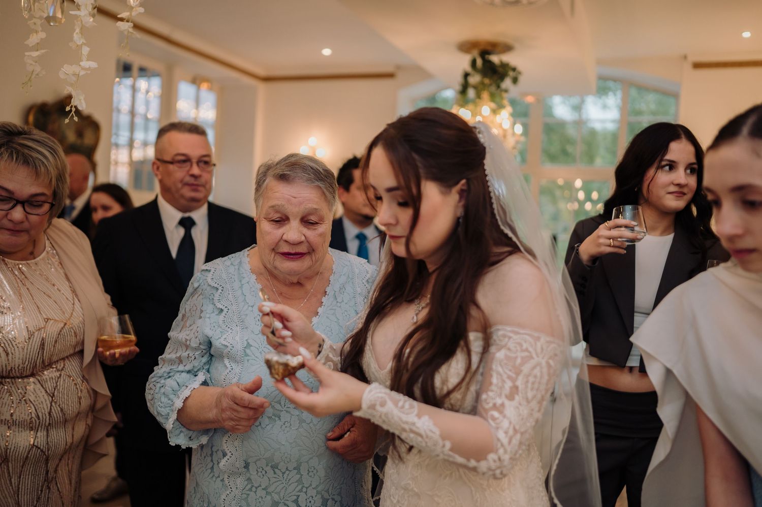 Bride in lace dress examining jewelry with older female guest at wedding reception.