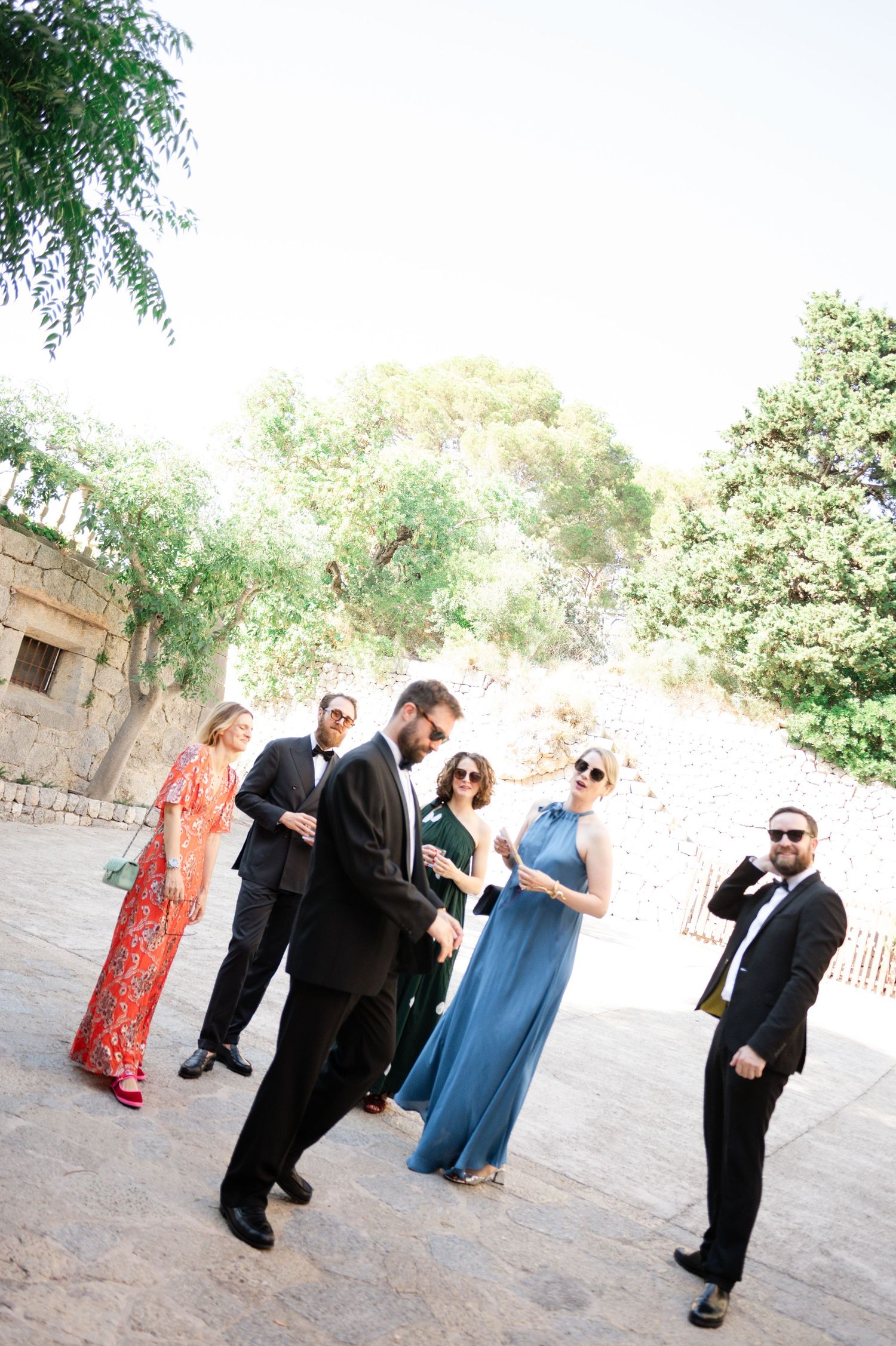 Wedding guests in formal attire socialize outdoors near stone walls and lush green trees on a sunny day.