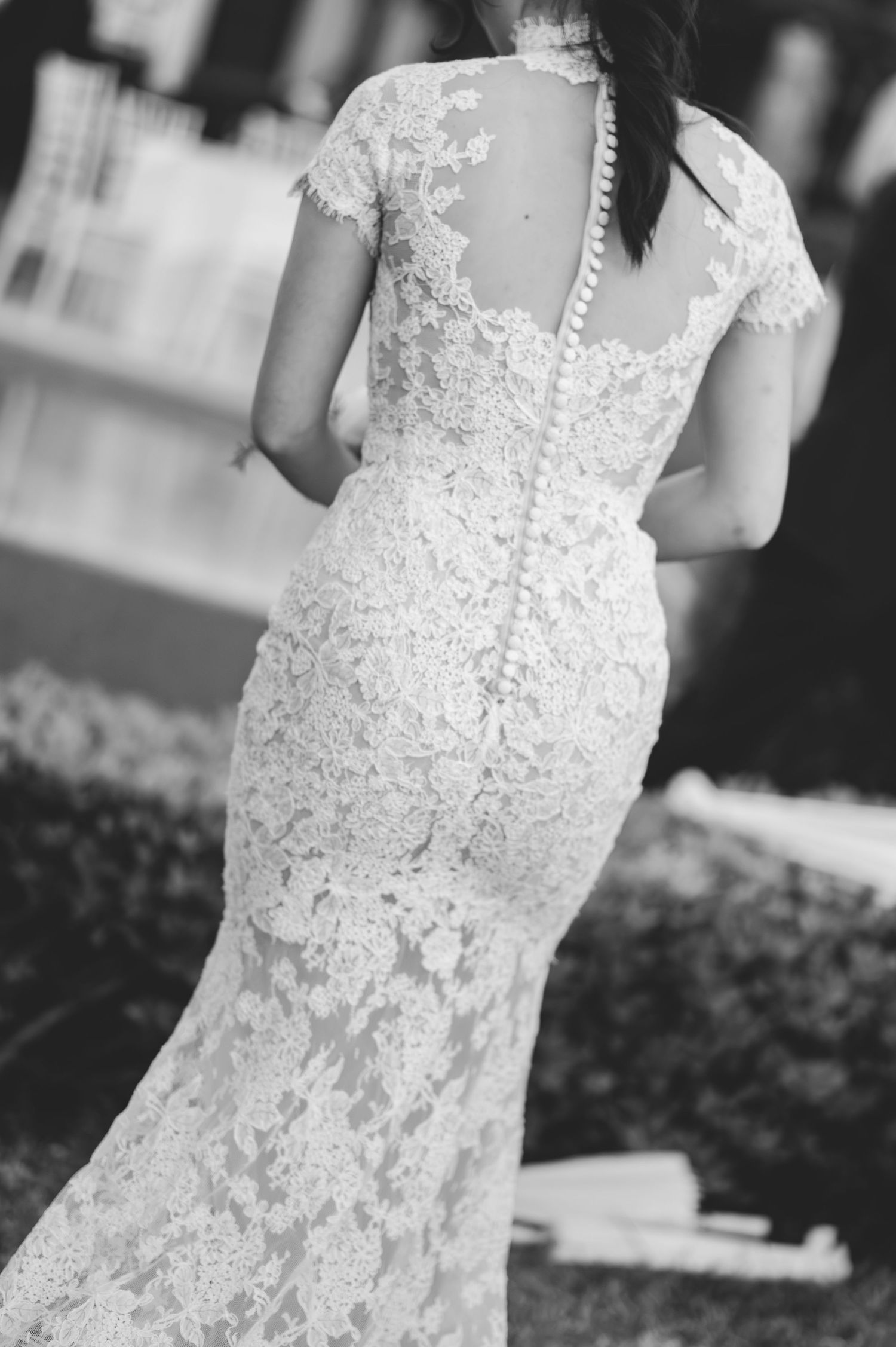 Elegant bride in a fitted lace wedding gown with cap sleeves and button-back detail, photographed from behind.