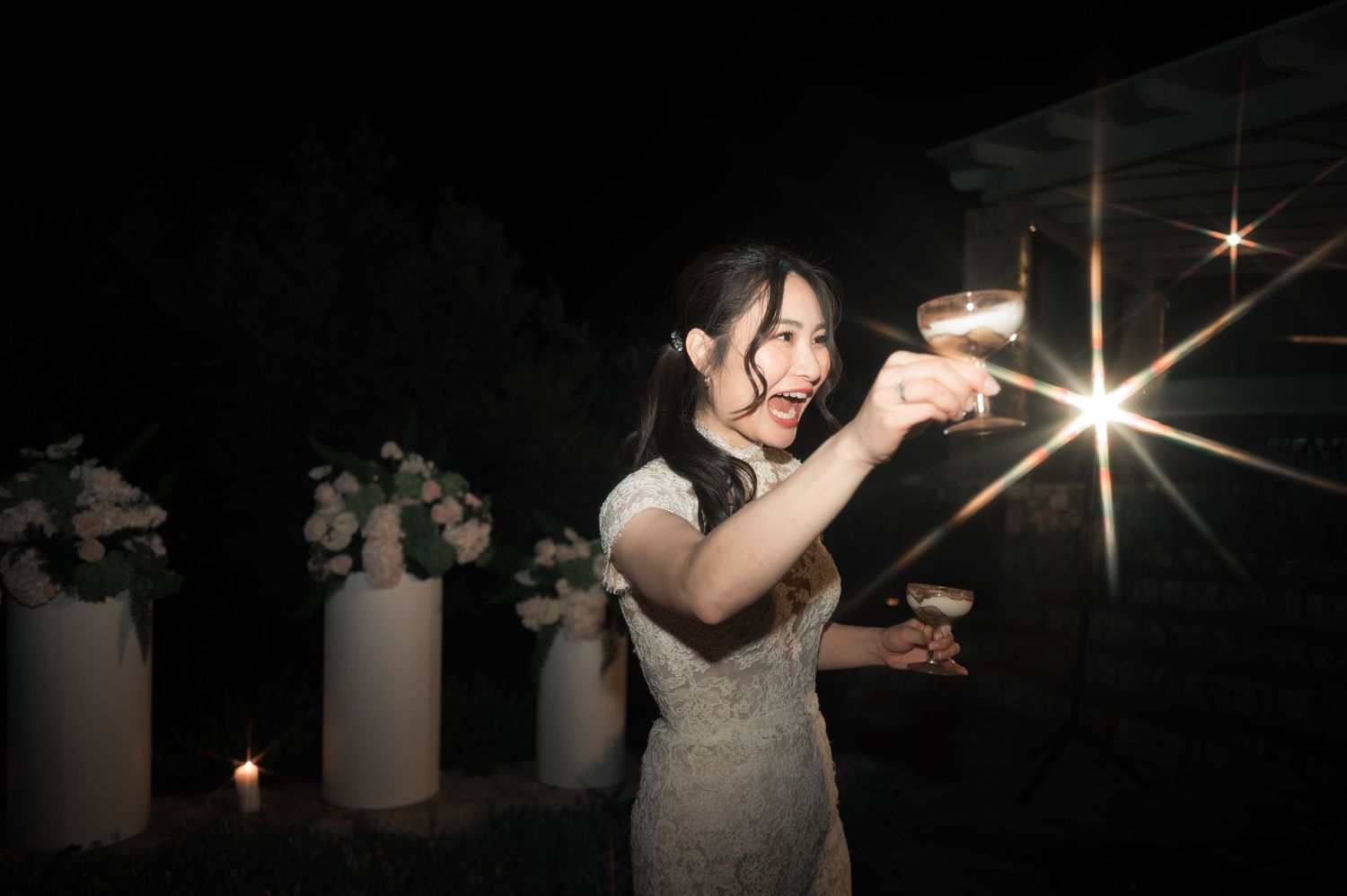 Bride in white lace dress holding a sparkler and champagne glass at a nighttime wedding.