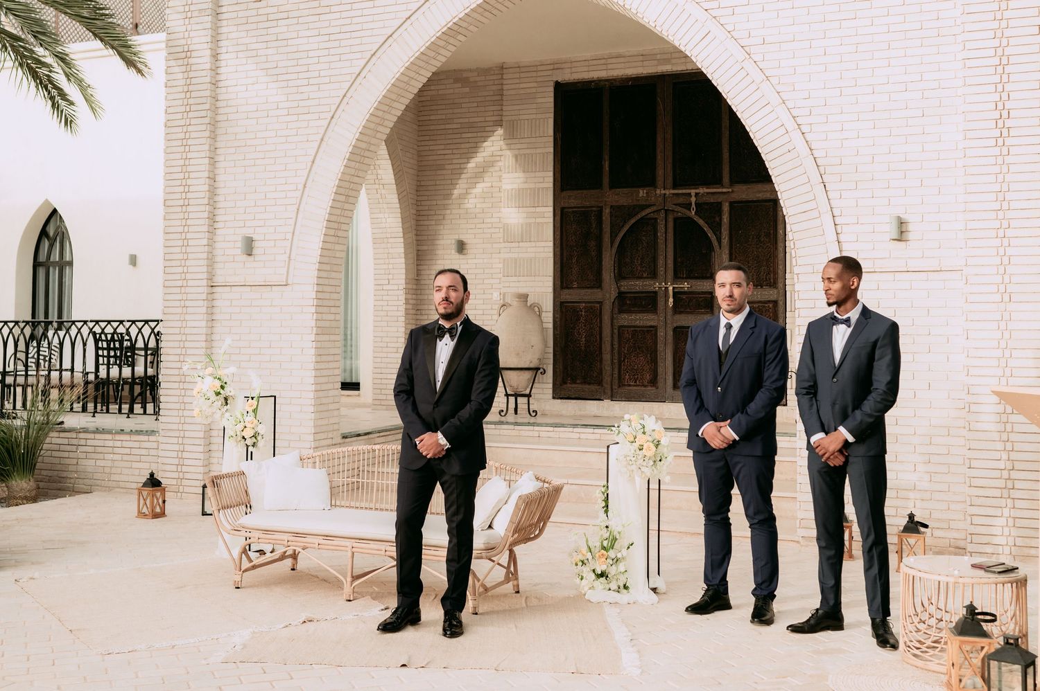 Three groomsmen in black tuxedos standing before an arched brick doorway decorated with white floral arrangements.