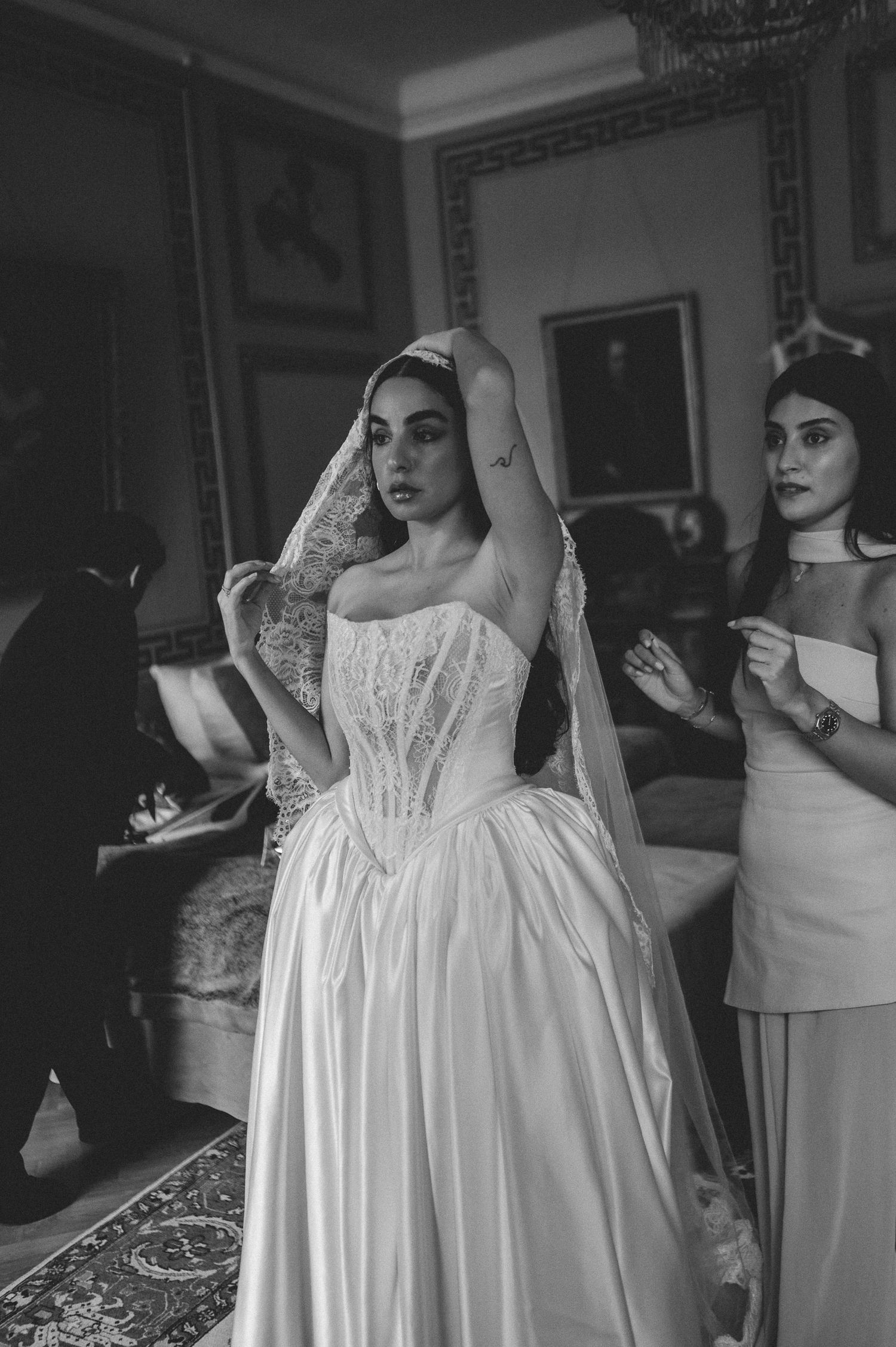 Black and white photo of bride in lace veil and corset gown being assisted by bridesmaids in elegant room.