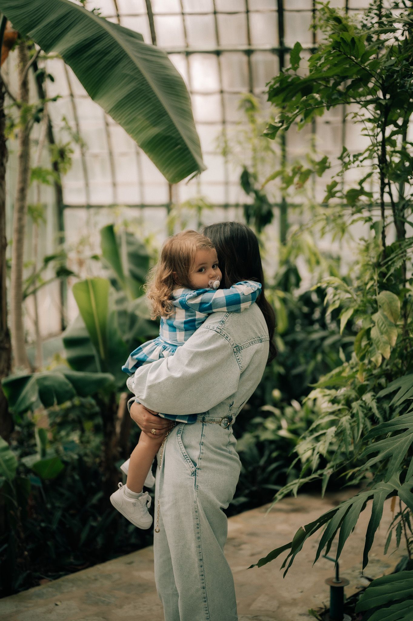 Mother tenderly holds her toddler in a lush tropical greenhouse surrounded by exotic plants.