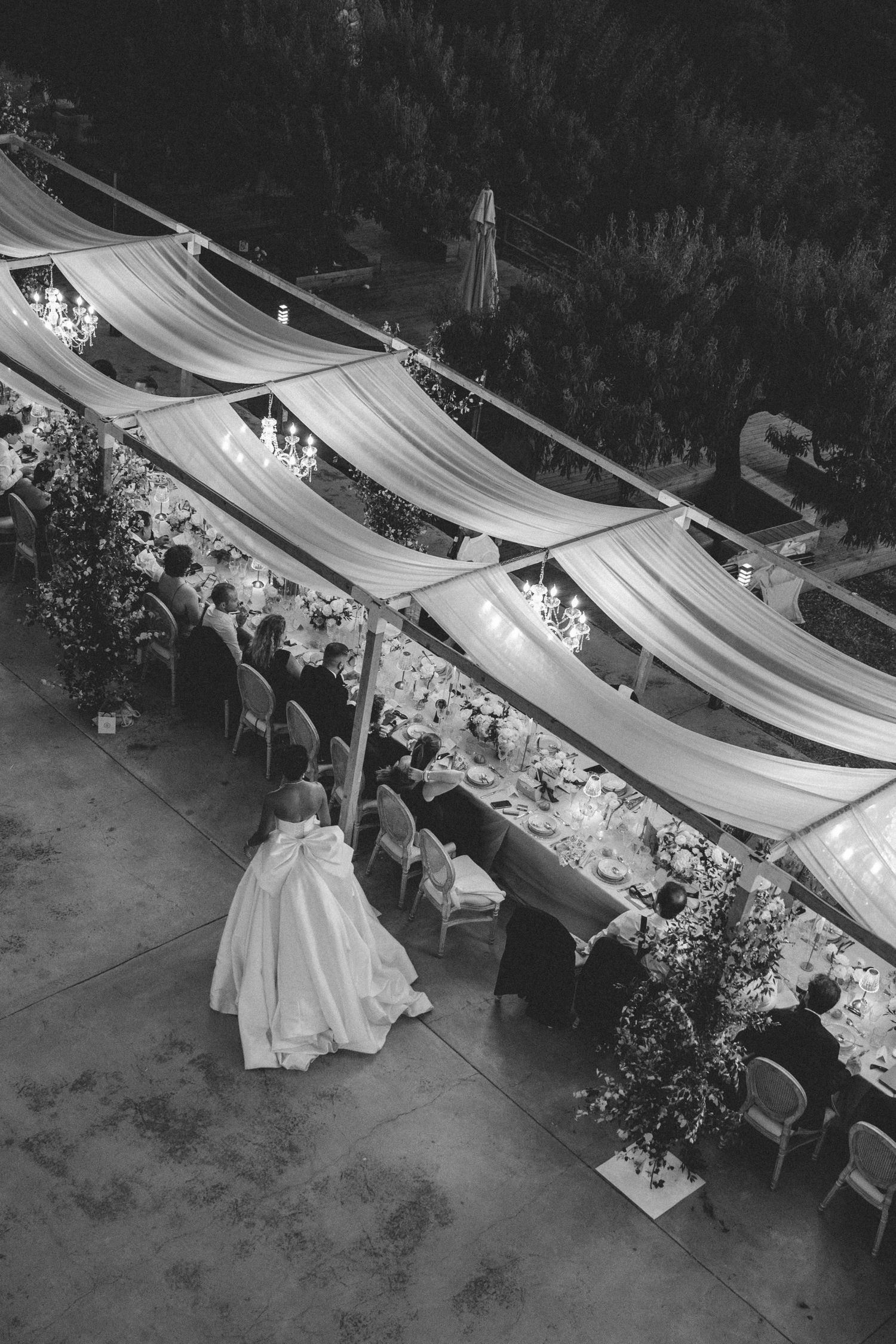 Aerial black and white view of bride in ball gown walking beside elegant draped wedding reception.