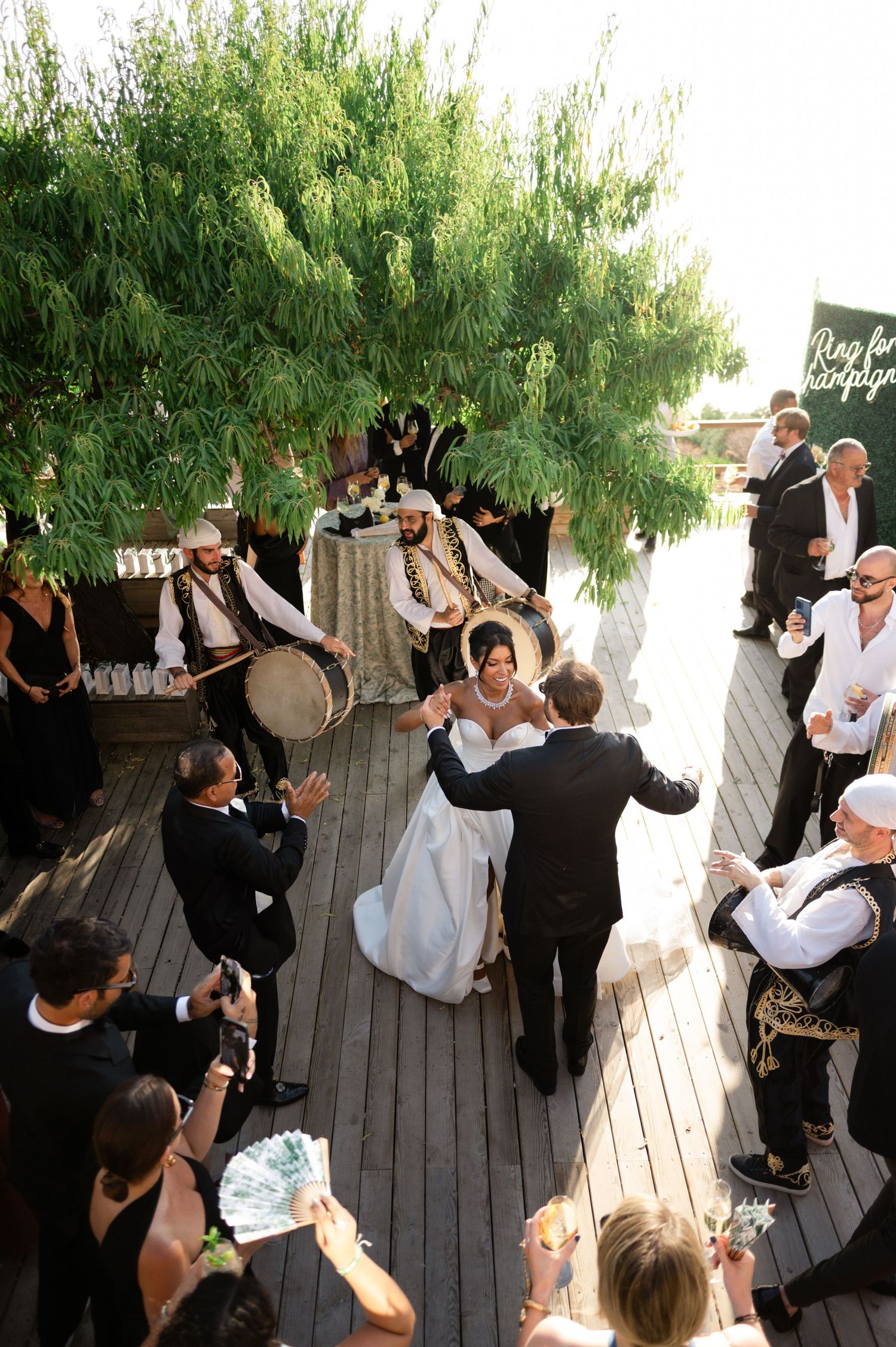 Bride and groom share first dance on outdoor wooden deck with traditional musicians and guests.