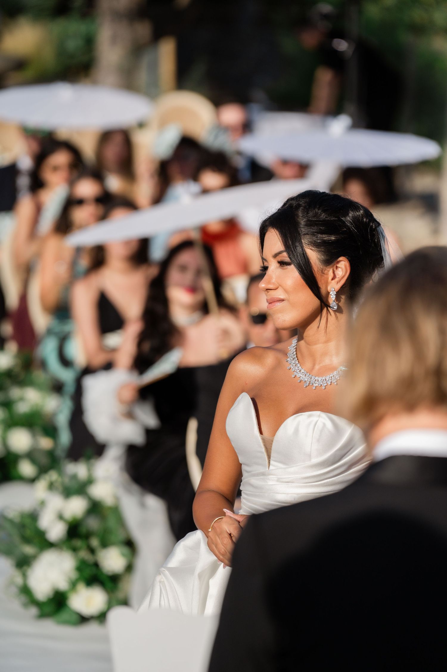 Beautiful bride in a strapless white gown with diamond necklace gazes at groom during outdoor ceremony.
