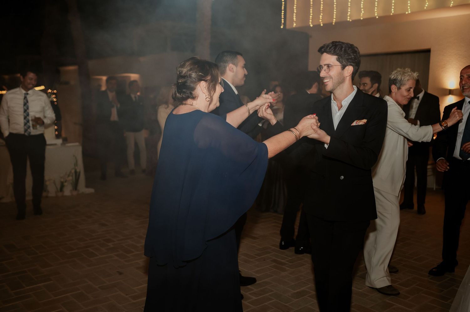 Guests dance together on outdoor wedding reception dance floor under atmospheric lighting.