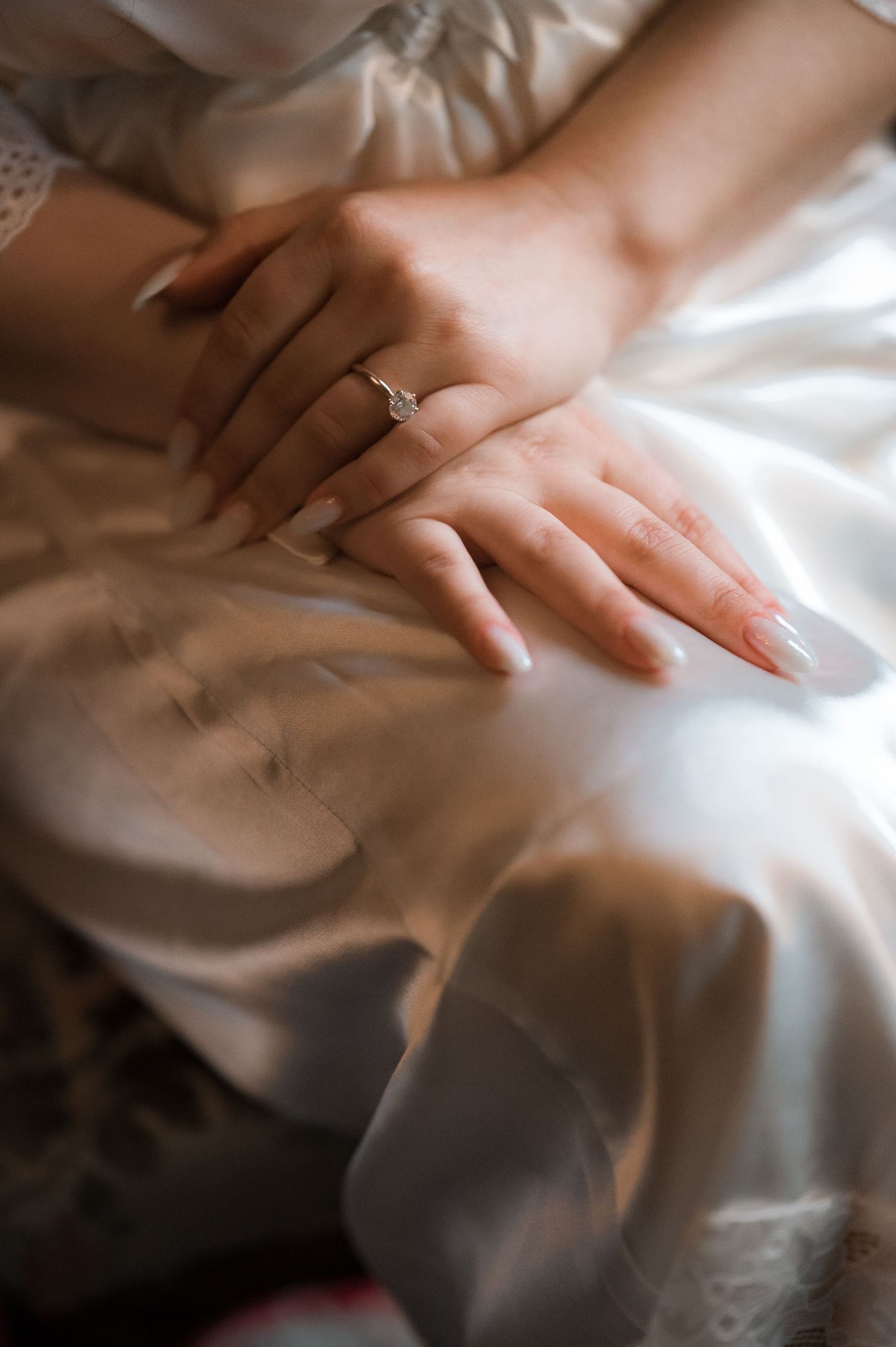 Close-up of bride\u2019s hands with a diamond engagement ring resting on her white satin gown.