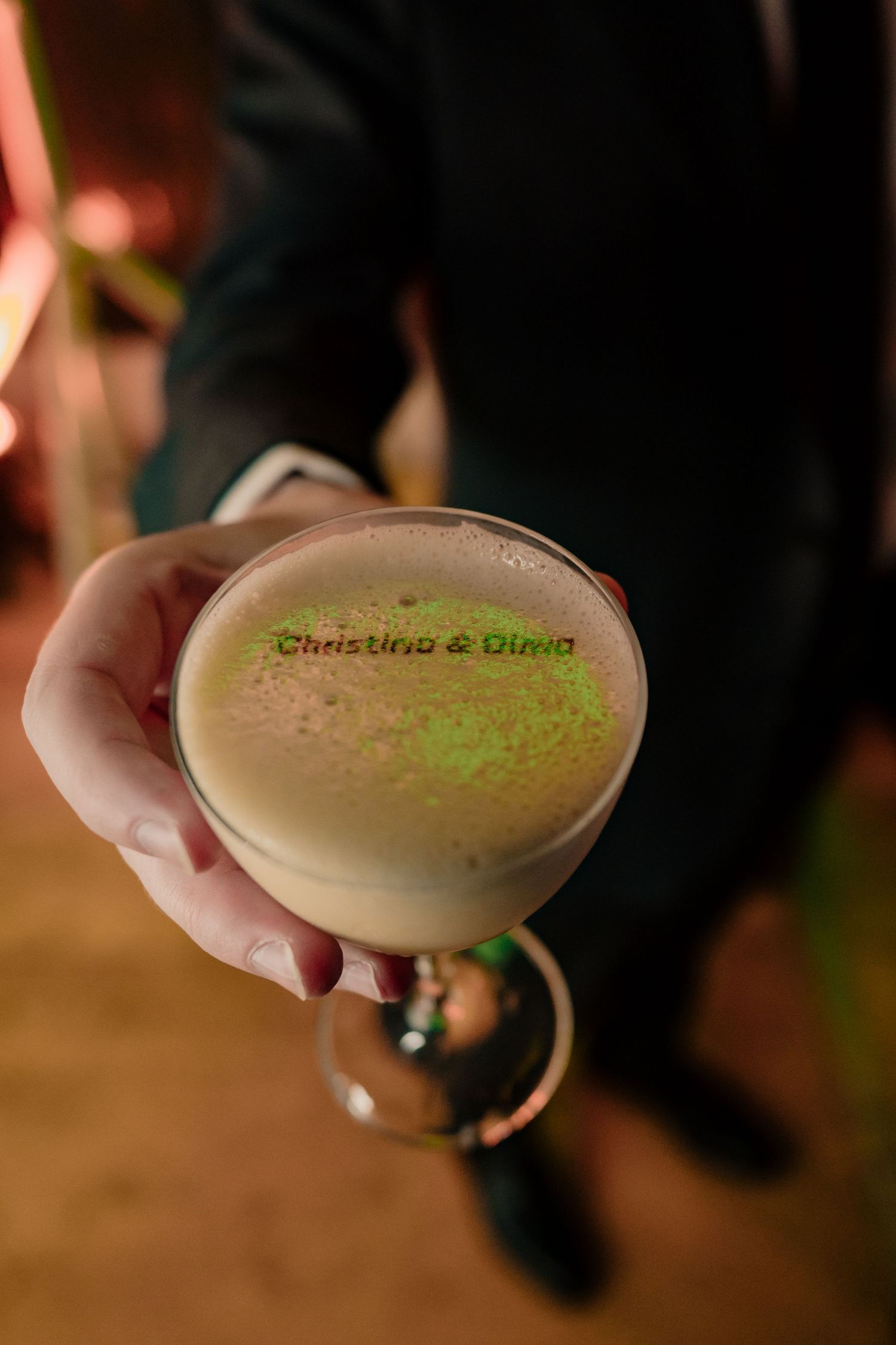 Hand holds cocktail with Christina and Dino laser-etched wedding logo on foam top.