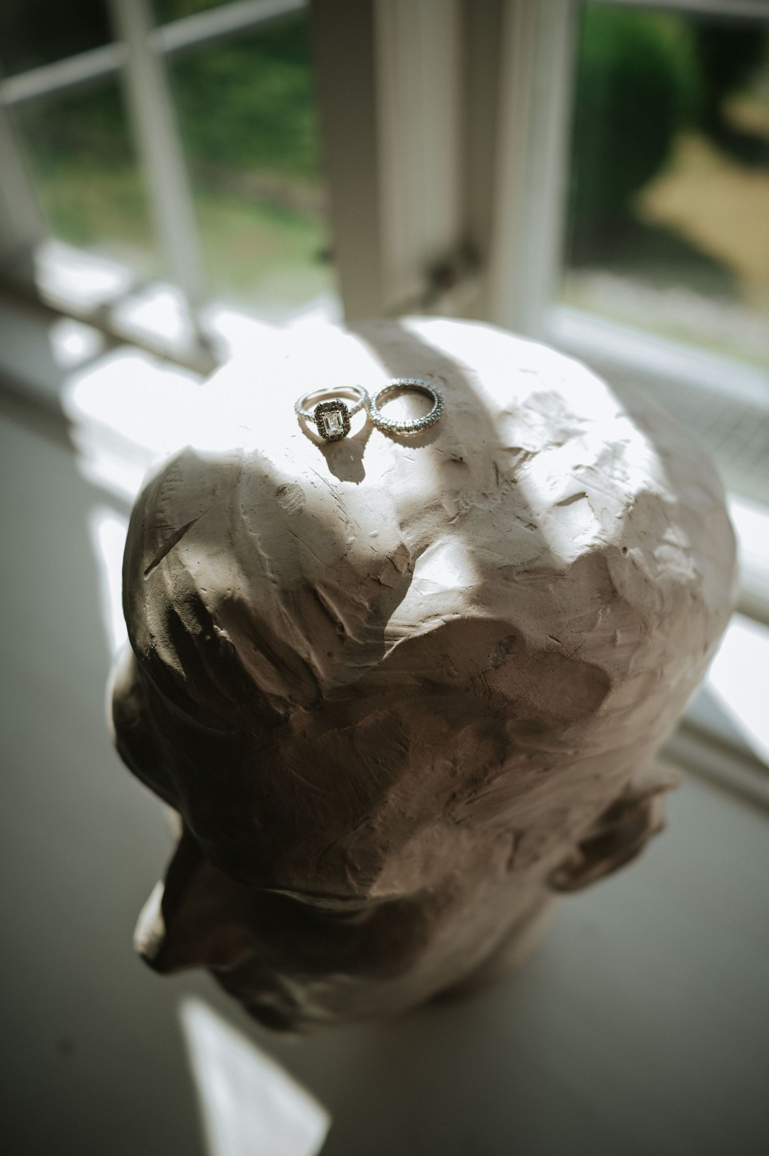 Two diamond wedding rings resting on top of a white plaster sculpture bust near window.