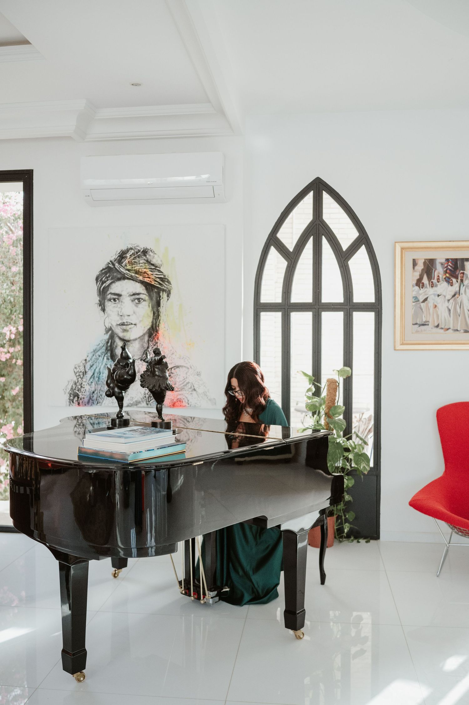 Woman in green dress playing grand piano in white room with large portrait artwork on wall.