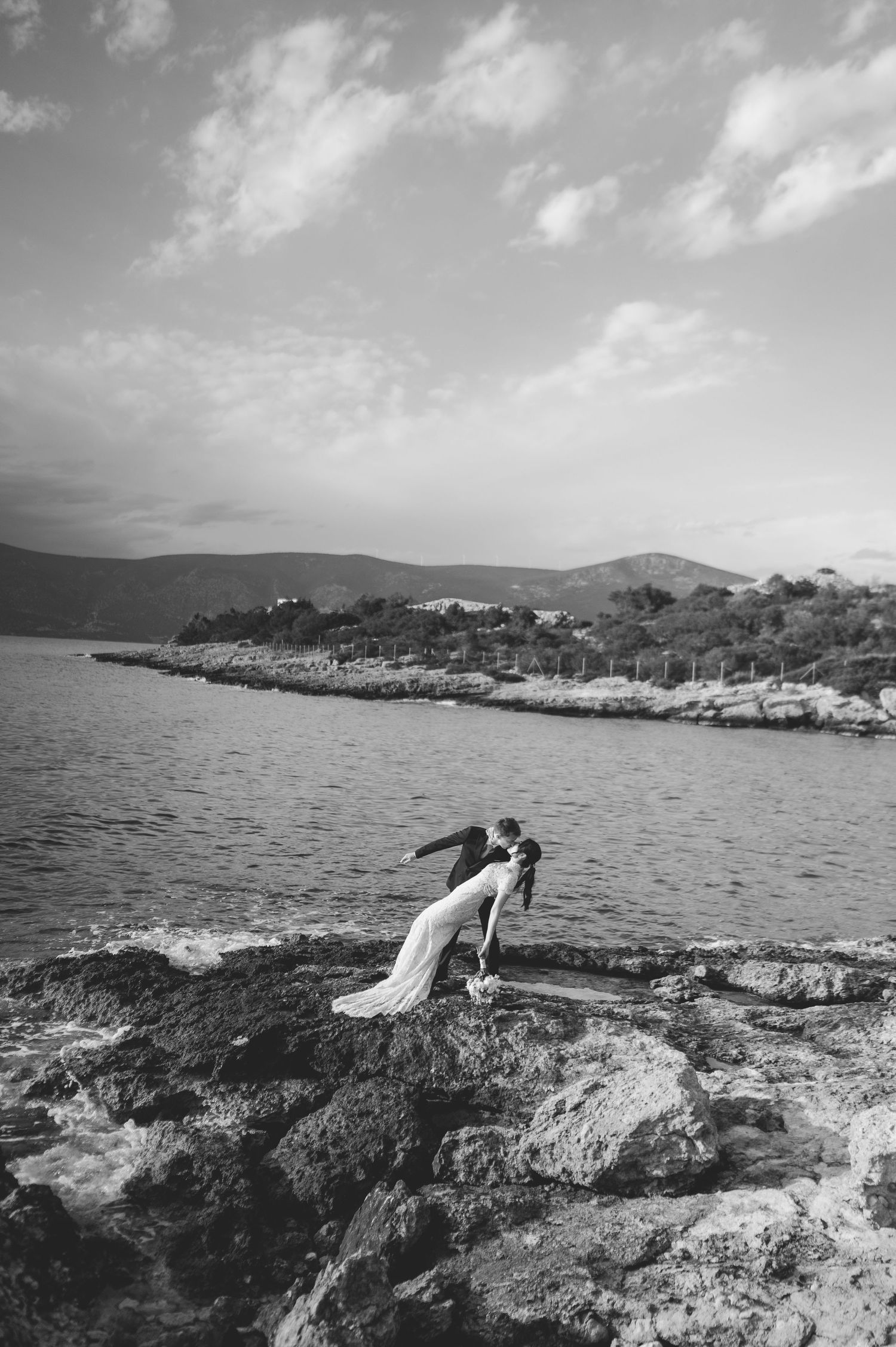 Bride and groom share romantic dip kiss on rocky shoreline in black and white photo.