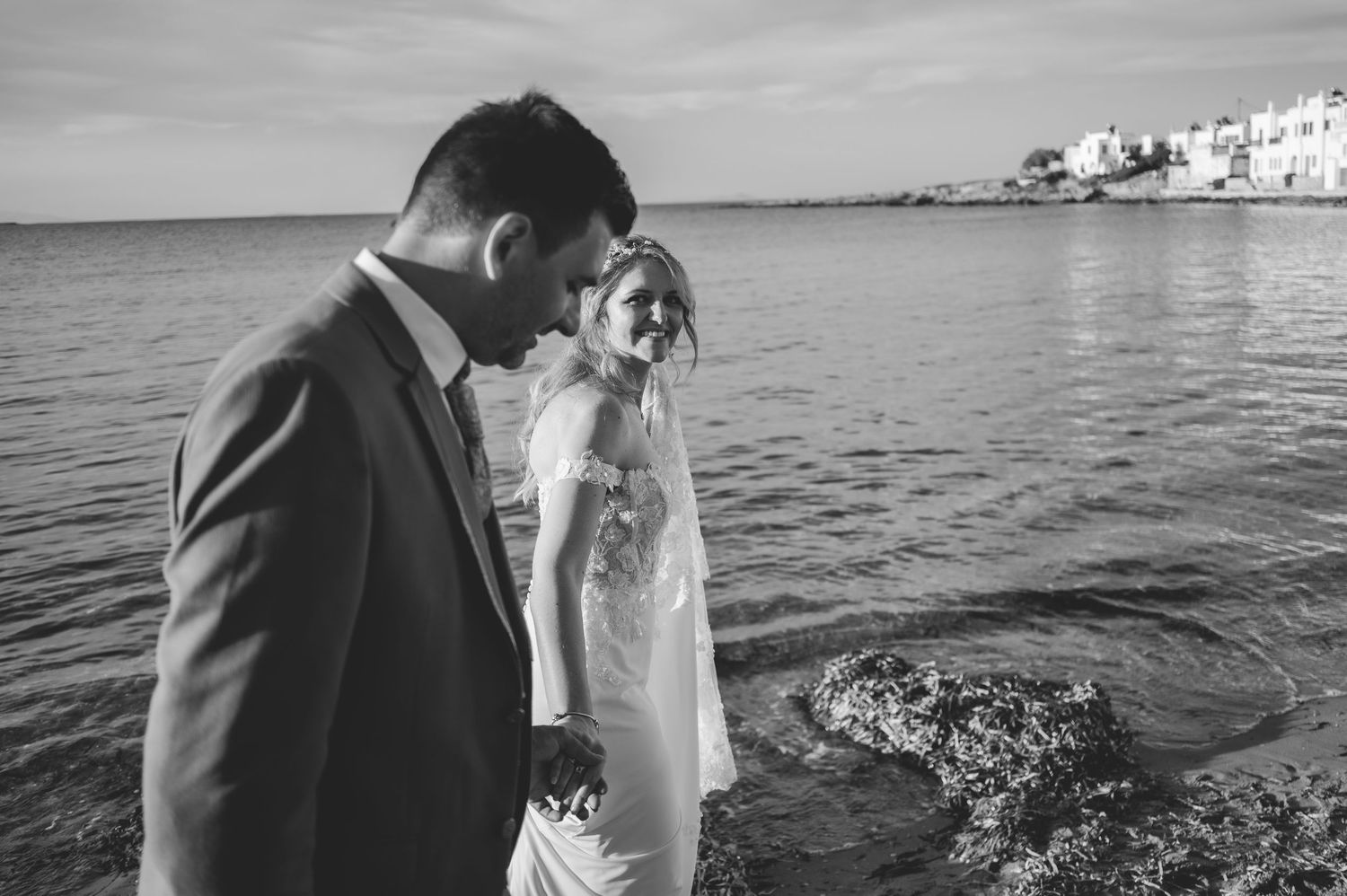 Newlyweds walk hand in hand along the shoreline of a Greek island in black and white.