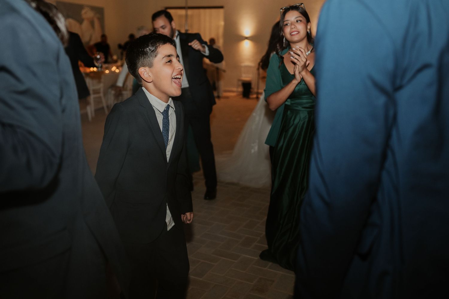 Young boy in suit smiles up at wedding guests while woman in green dress claps during reception celebration.