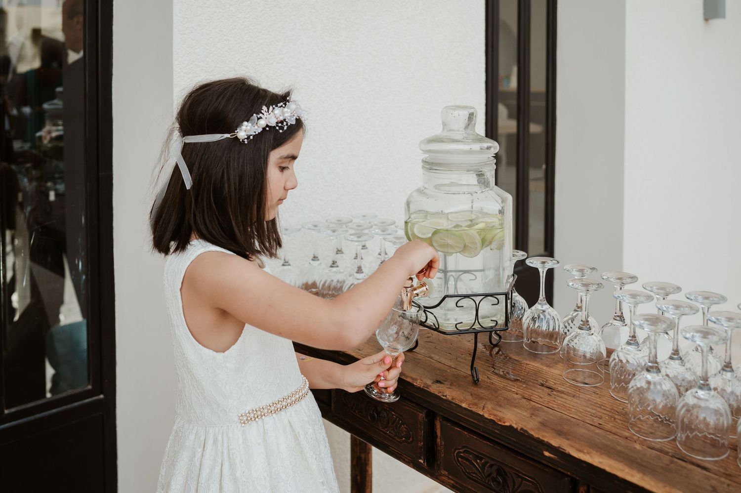 Young flower girl in white dress reaching for glassware at elegant wedding drink station.